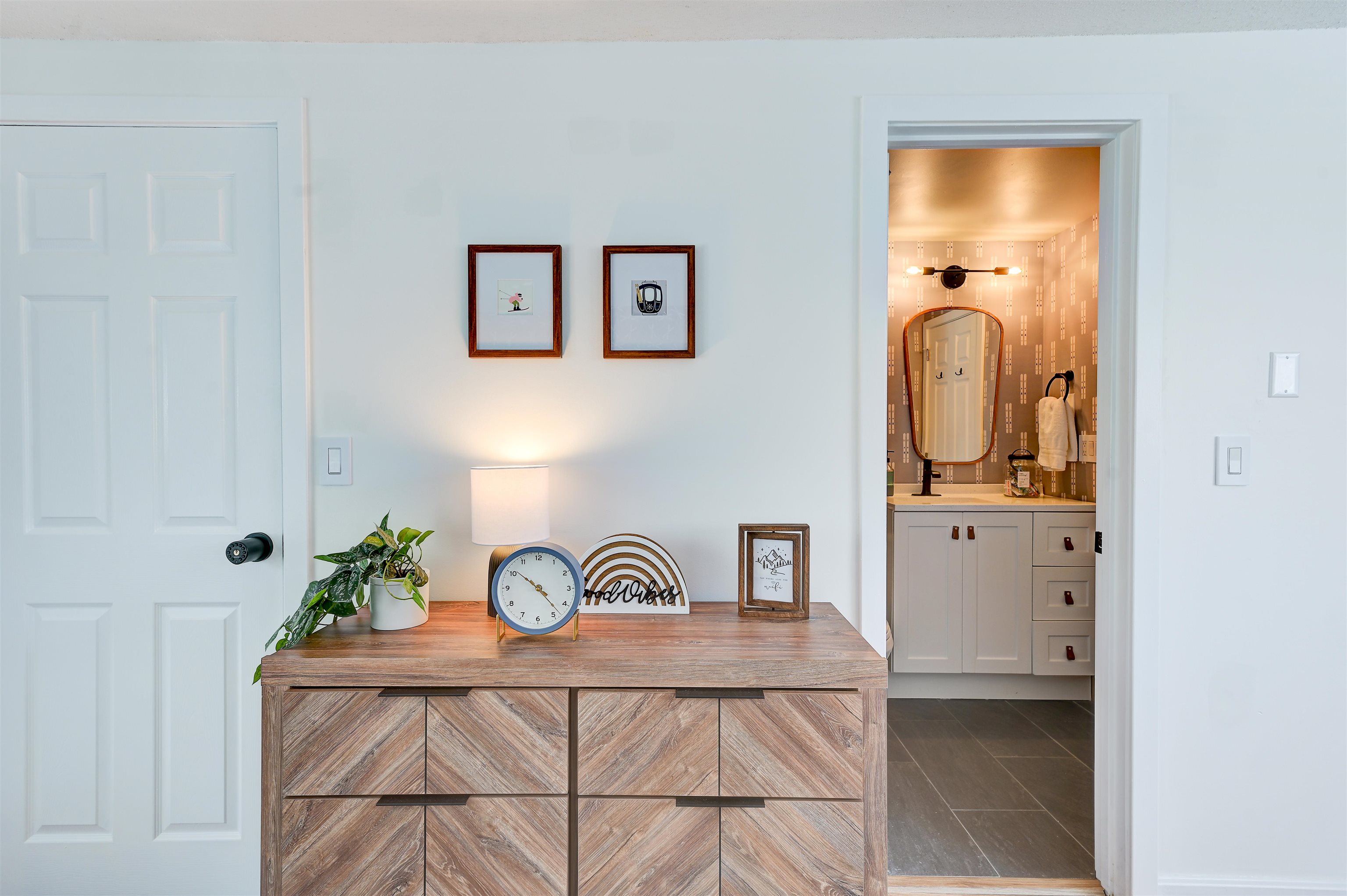 Entryway with a wood-grain console and clock, looking into a bathroom with patterned wallpaper and a vanity.