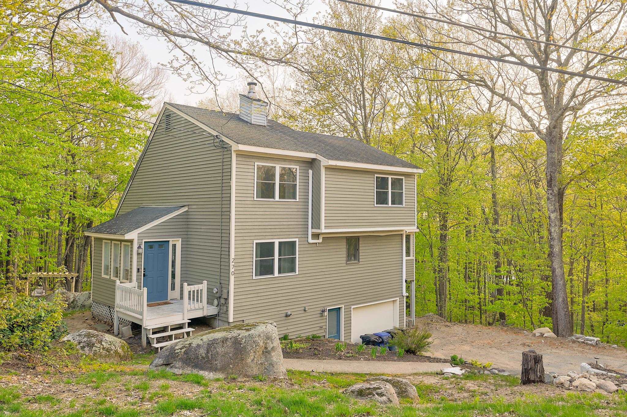 Two-story gray-green home with a blue front door, small porch, and rocky wooded yard.