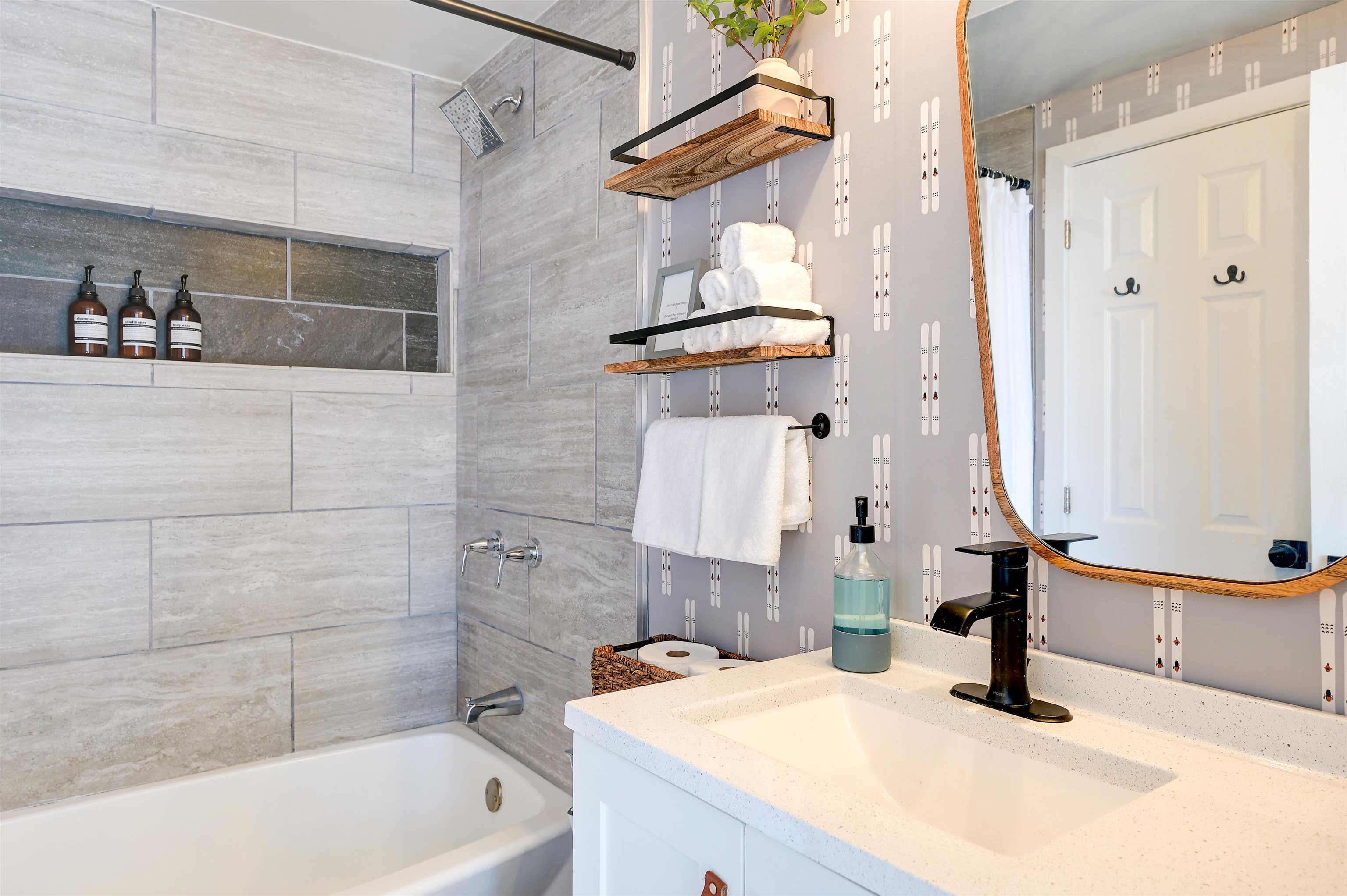 Bathroom with gray tiled tub surround, patterned wallpaper, and a white vanity with a black faucet.