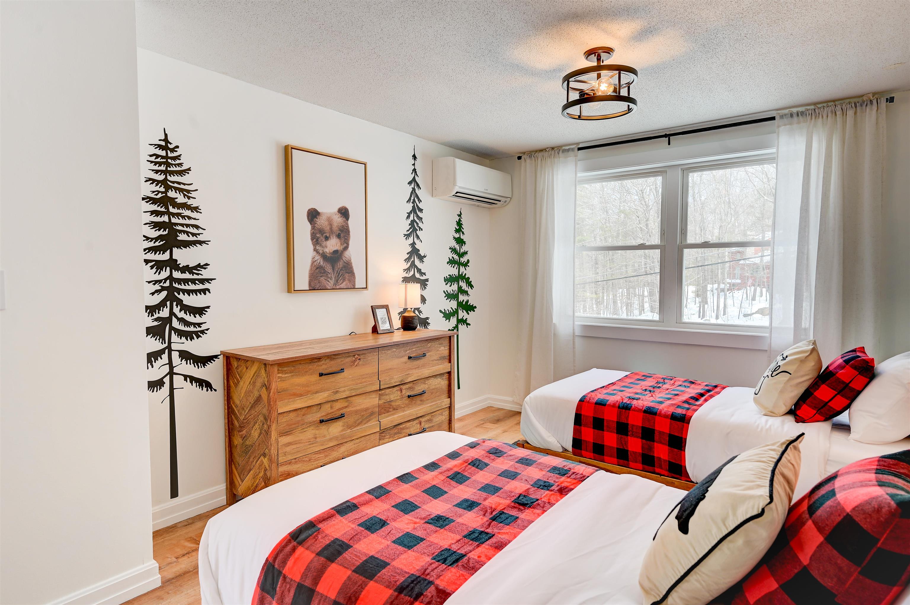 Bedroom with two plaid-covered beds, a rustic dresser, and a window overlooking snowy woods.