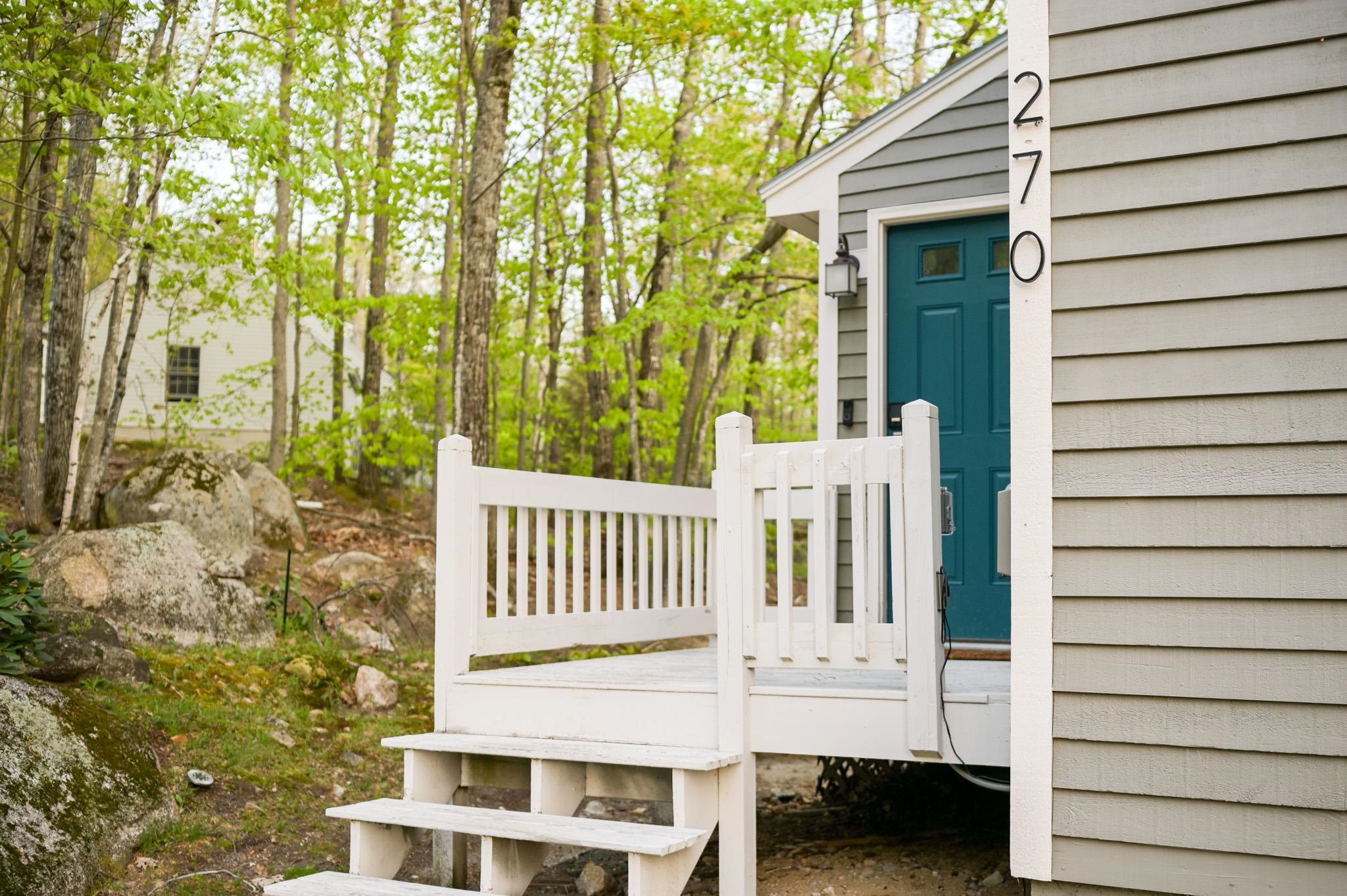 Home entrance with a teal door, white porch, and wooded, rocky yard.