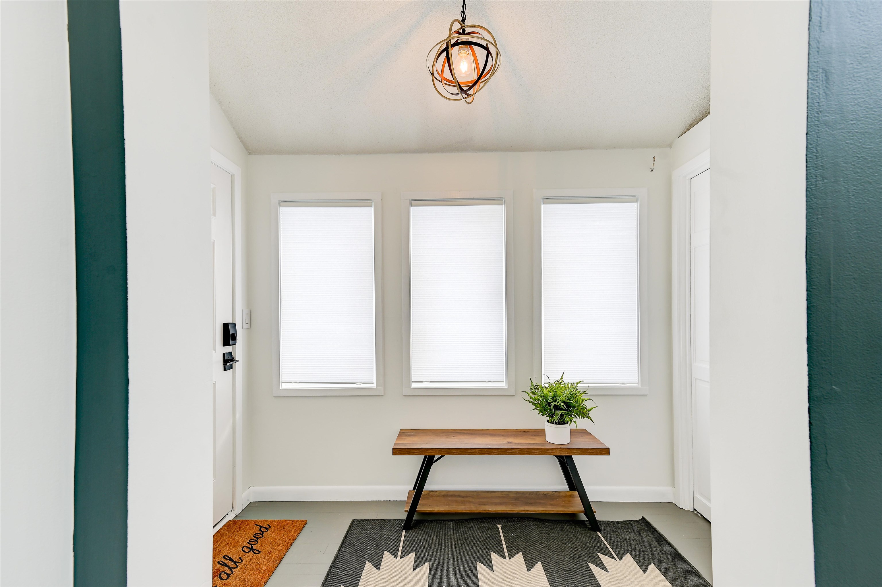 Entryway with three white-blinded windows, a wood console table, potted plant, and geometric pendant light.