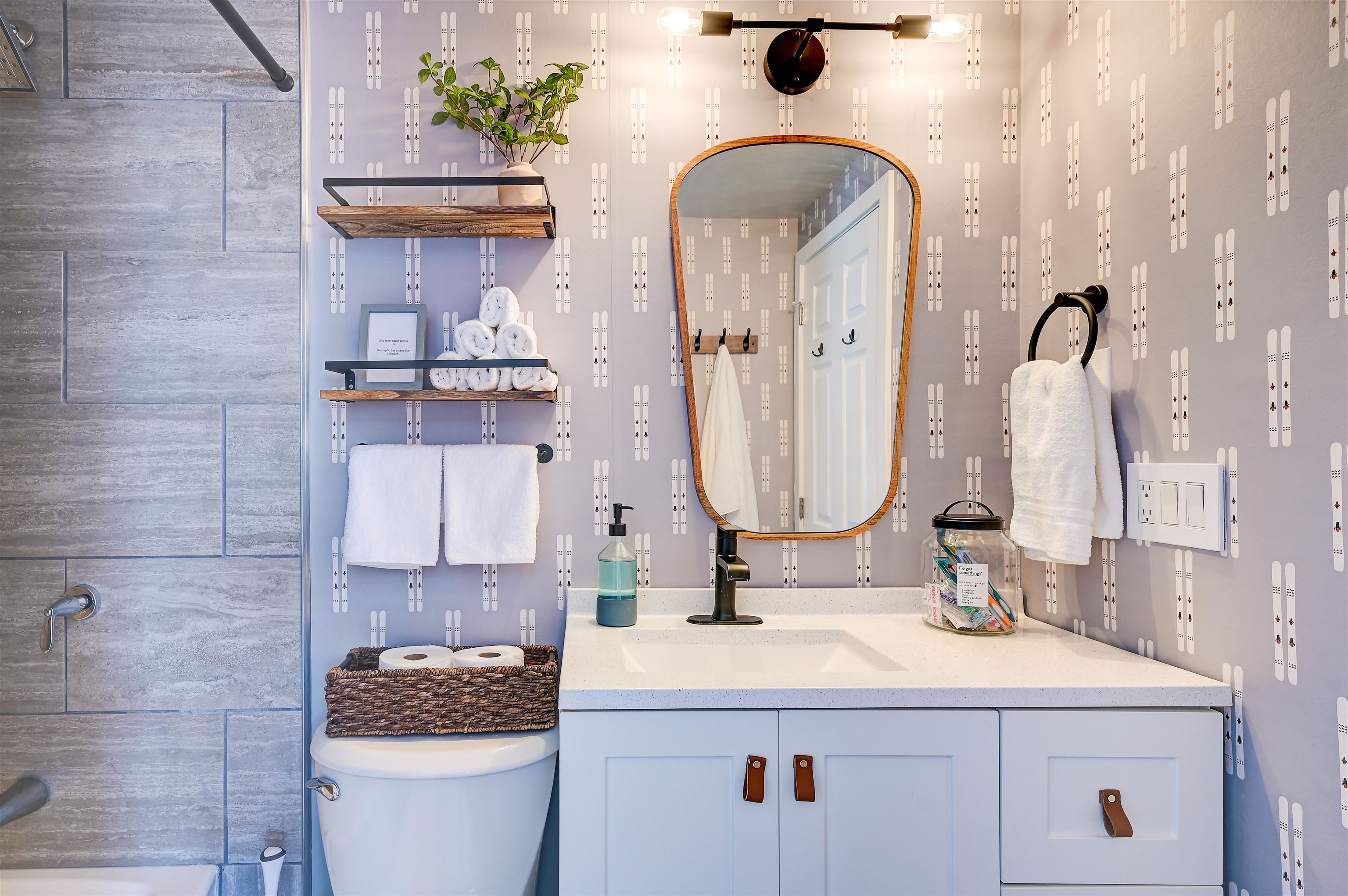 Bathroom vanity with a wood-framed mirror, patterned wallpaper, and gray stone-tiled shower.