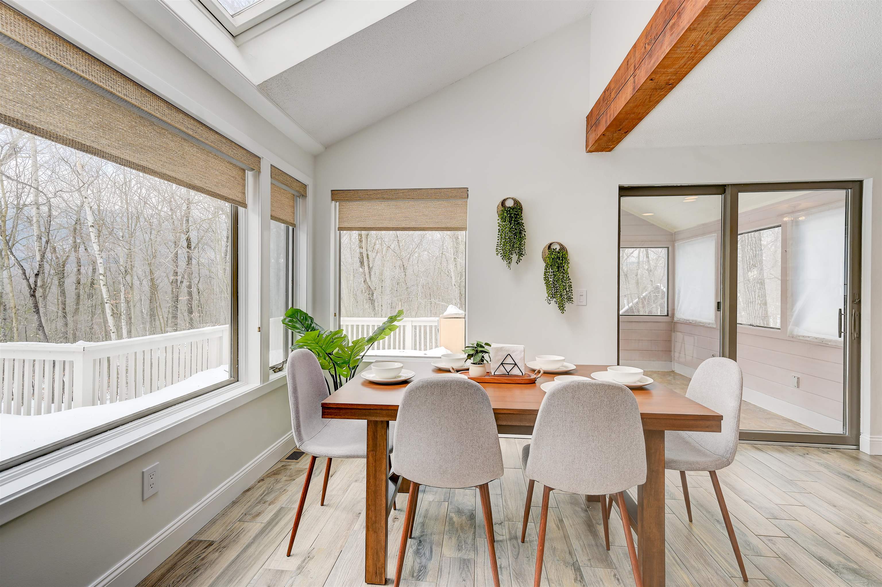 Dining room with a wood table, large windows overlooking snowy woods, and a sliding door to an enclosed porch.