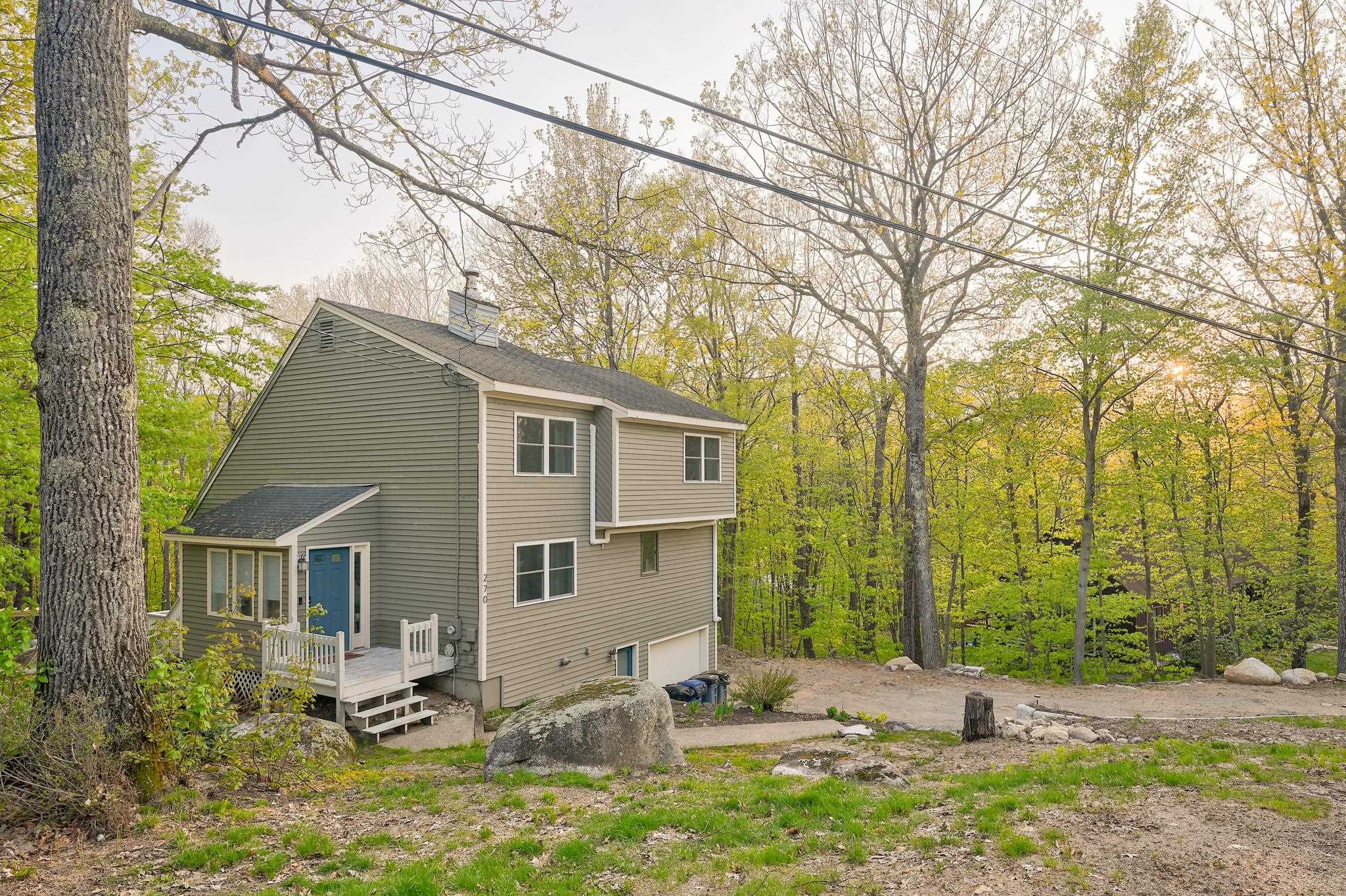 Two-story gray house with a blue door and small deck, surrounded by autumn trees.