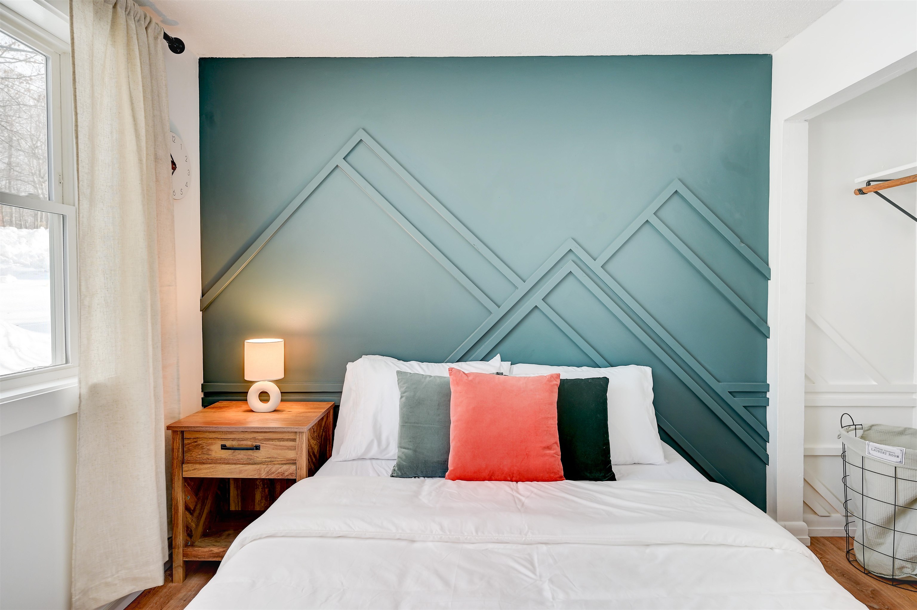 Bedroom with a teal geometric accent wall, white bedding, coral pillow, and wooden bedside table.