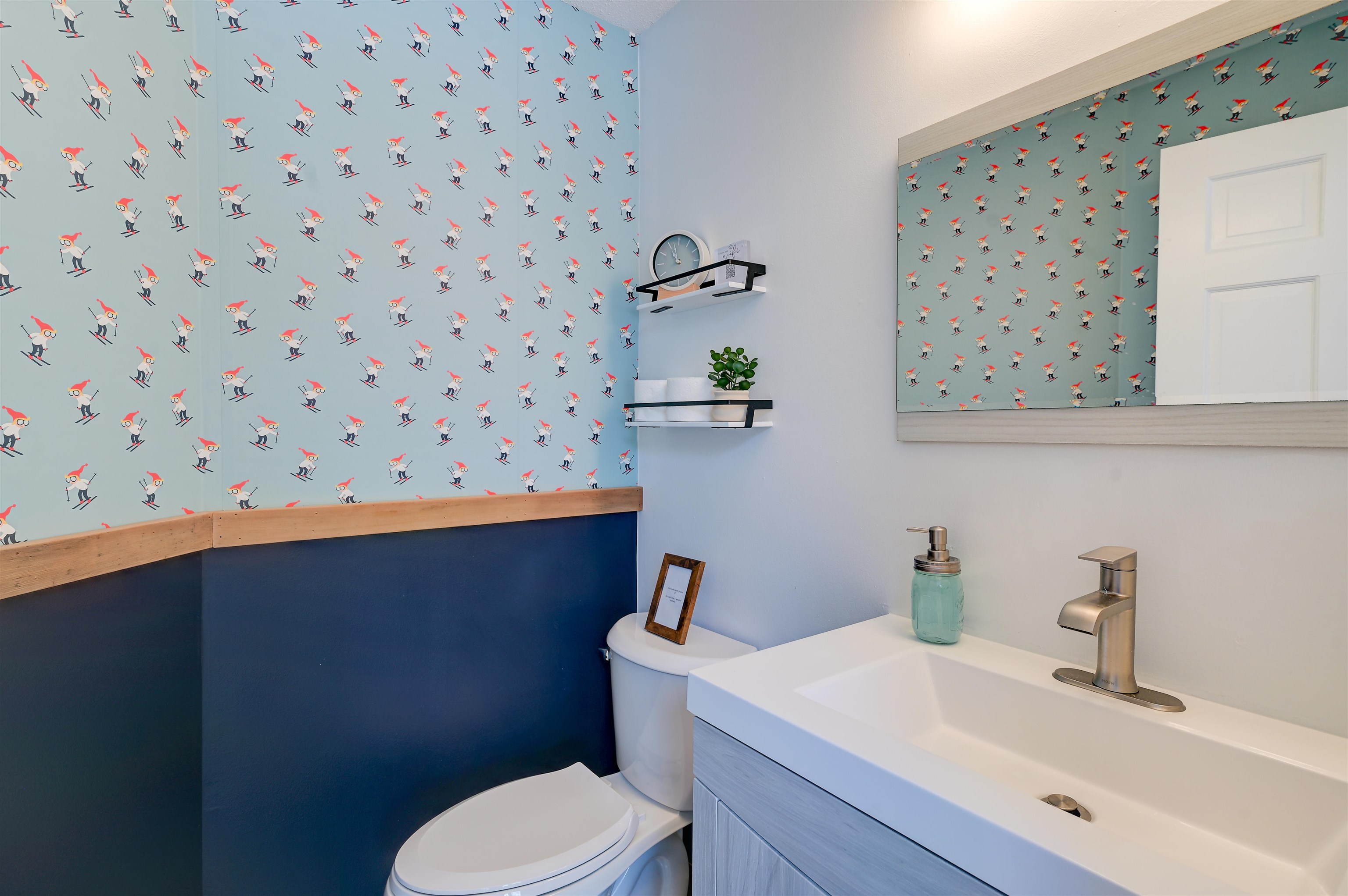 Bathroom with skier-patterned wallpaper, blue wainscoting, white vanity, and toilet.