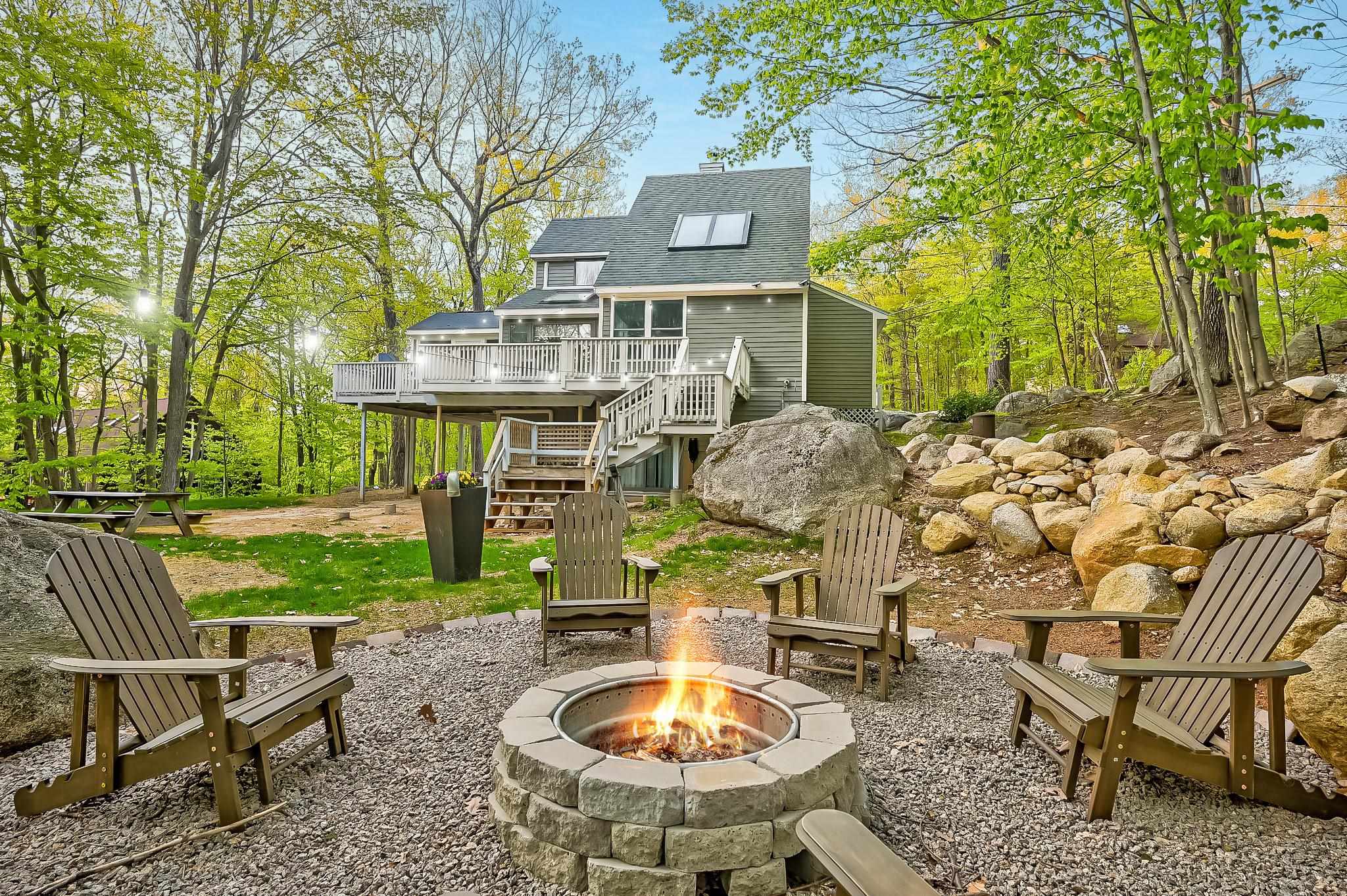Wooded home with an elevated deck and a stone fire pit surrounded by Adirondack chairs.