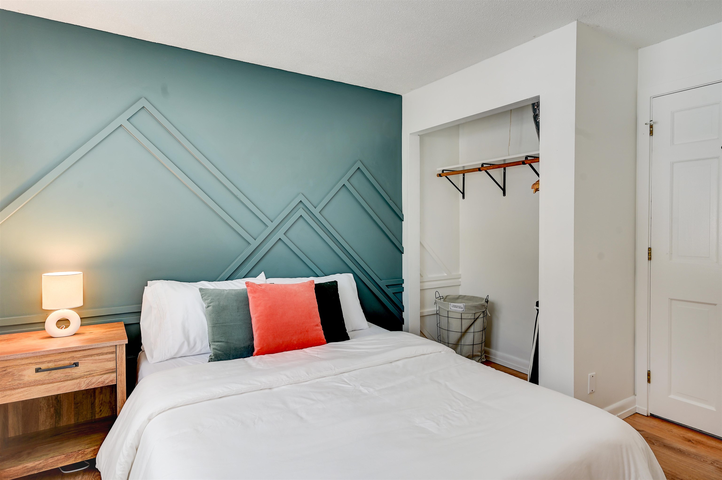 Staged bedroom with a white bed, geometric teal accent wall, and open closet nook.