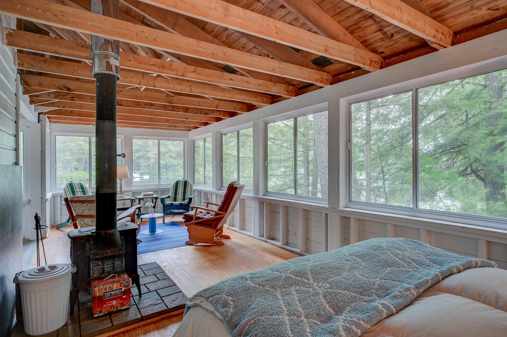 Sunroom with a bed, exposed timber ceiling, wood stove, and wide windows overlooking trees.