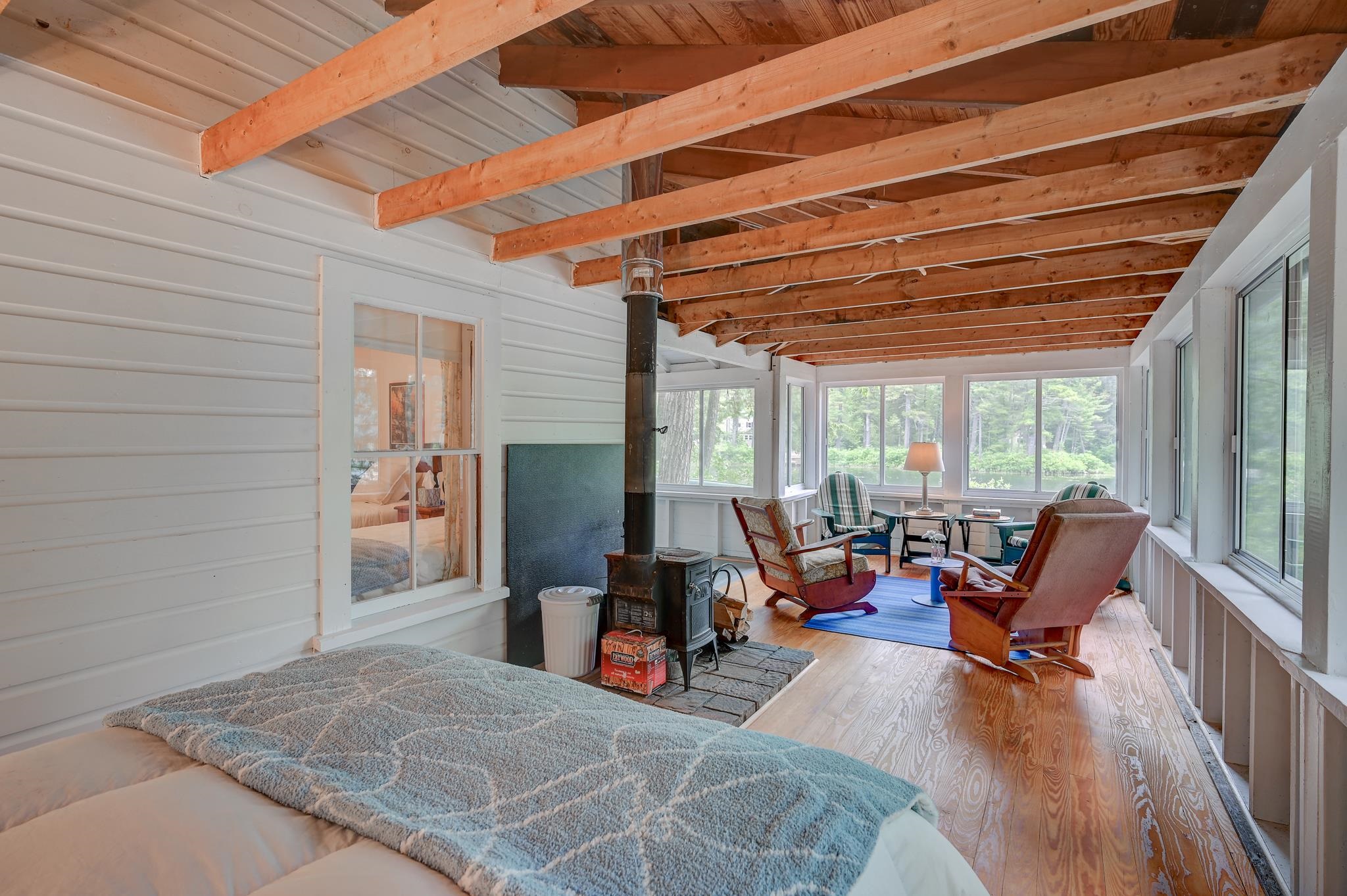Furnished sunroom with exposed beams, a wood stove, and broad windows overlooking water.