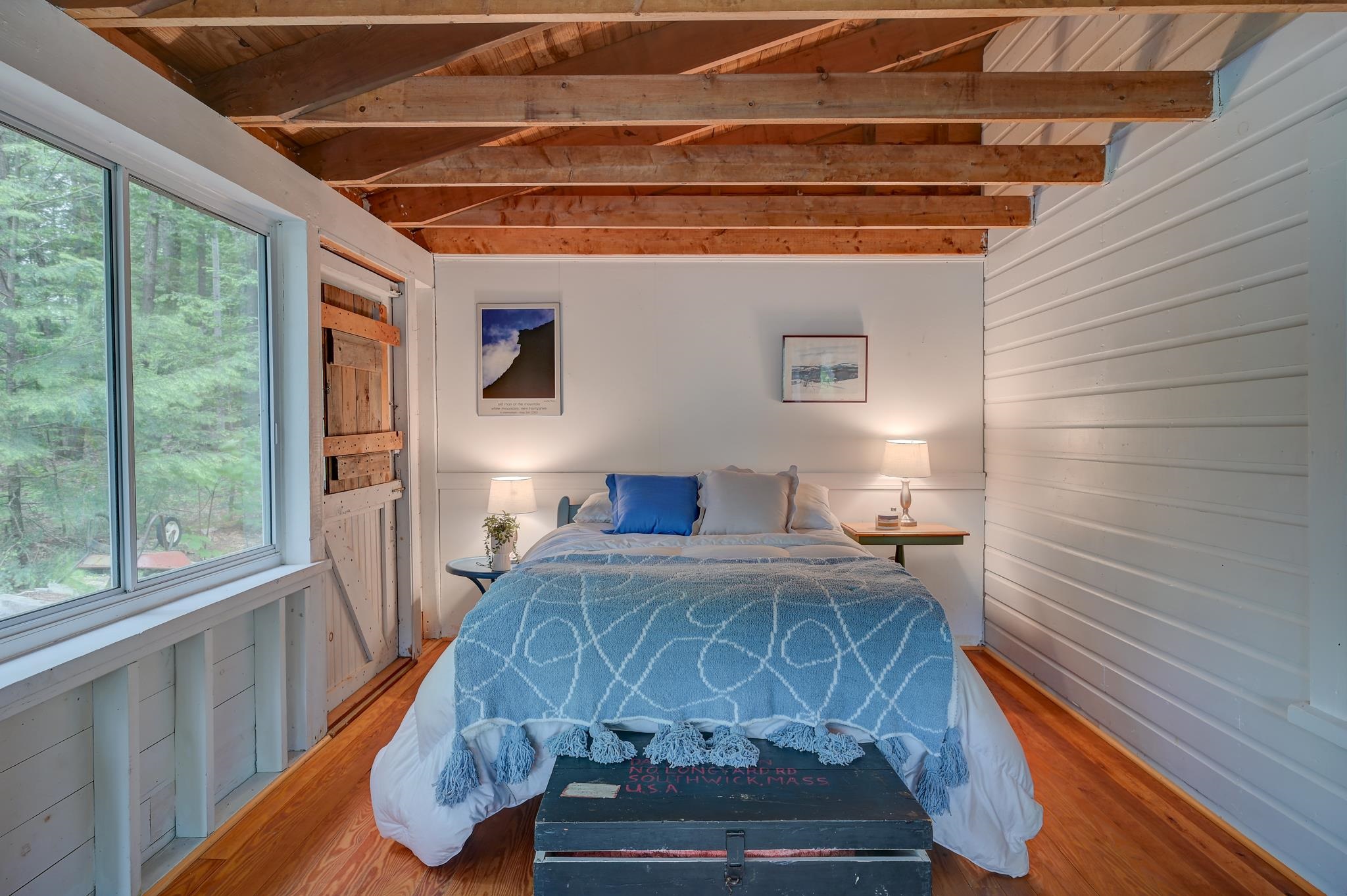 Bedroom with blue bedding, exposed ceiling beams, hardwood floors, and a large window overlooking trees.