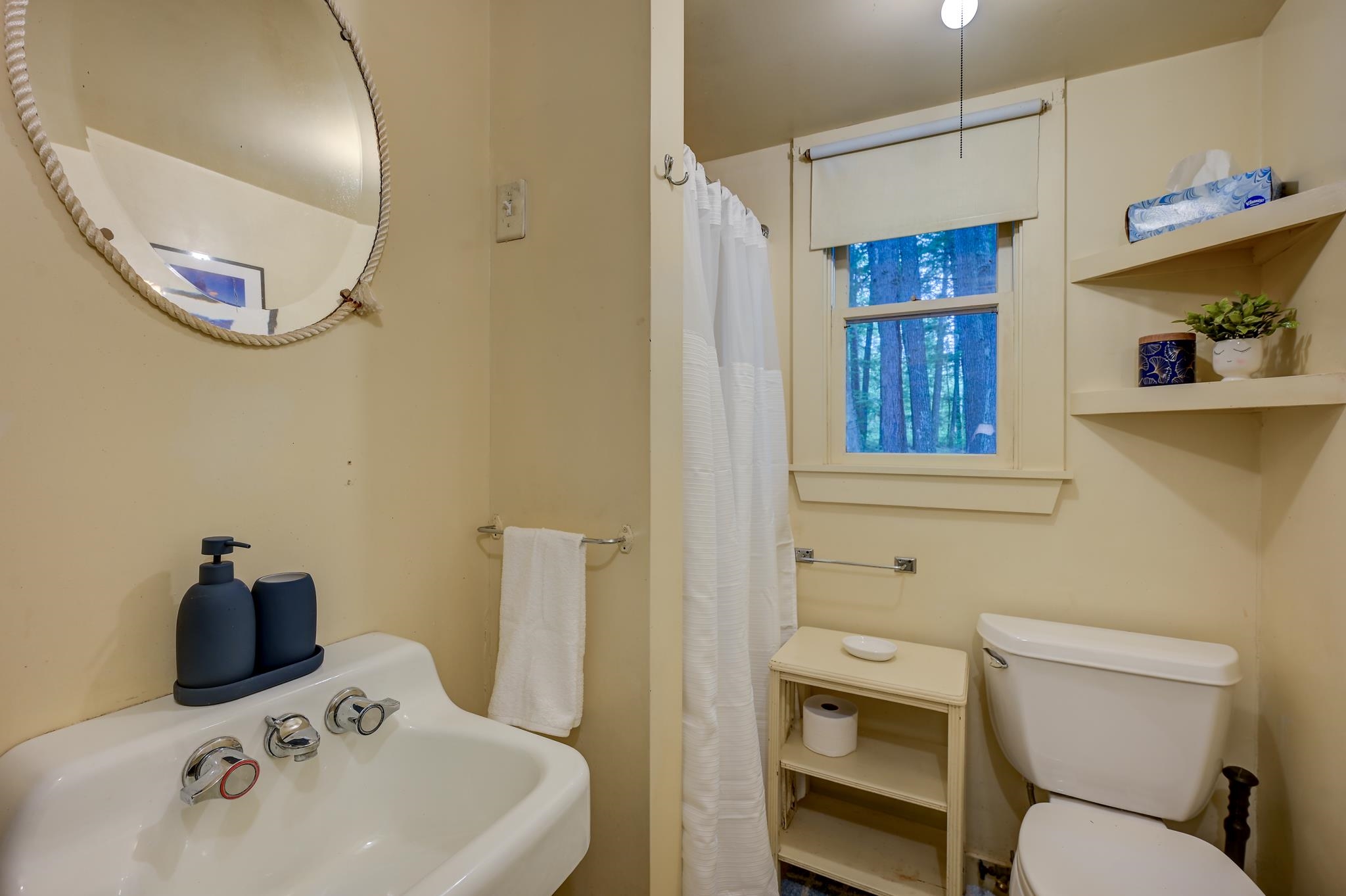 Bathroom with rope-framed mirror, white shower curtain, and window overlooking trees.