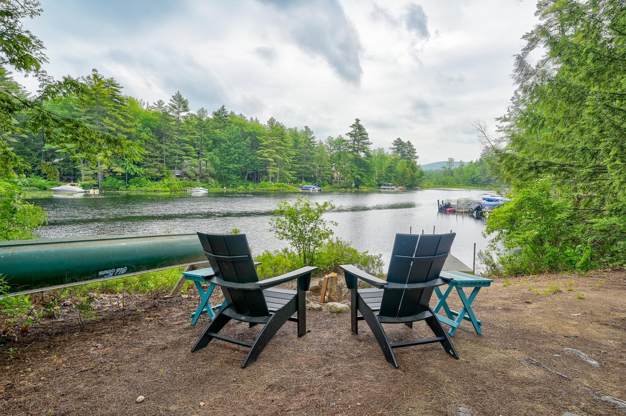 Two Adirondack chairs overlook a wooded lake, with a canoe beside them and boats across the water.