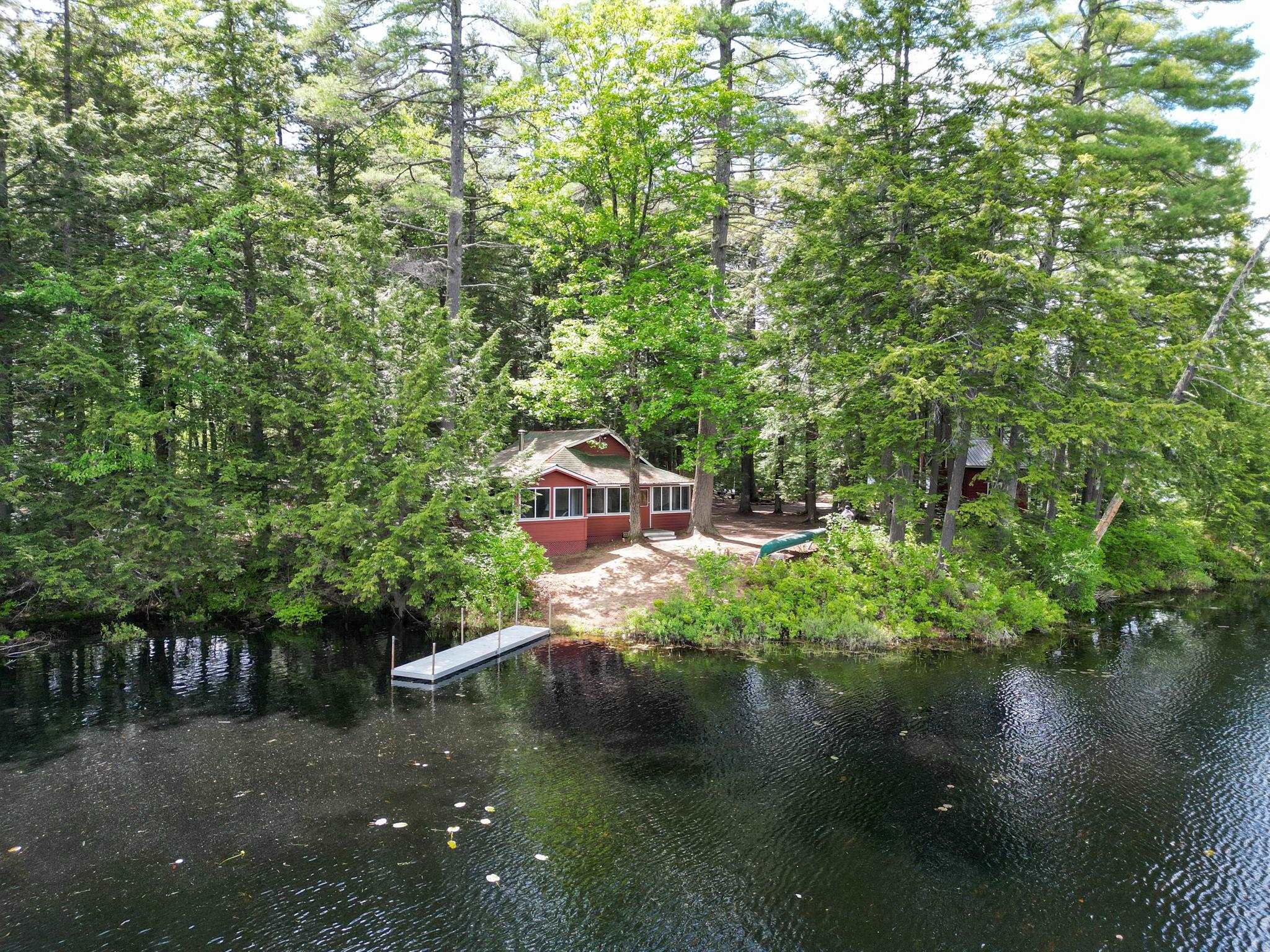 Red cabin among trees beside a lake, with a small dock extending into the water.