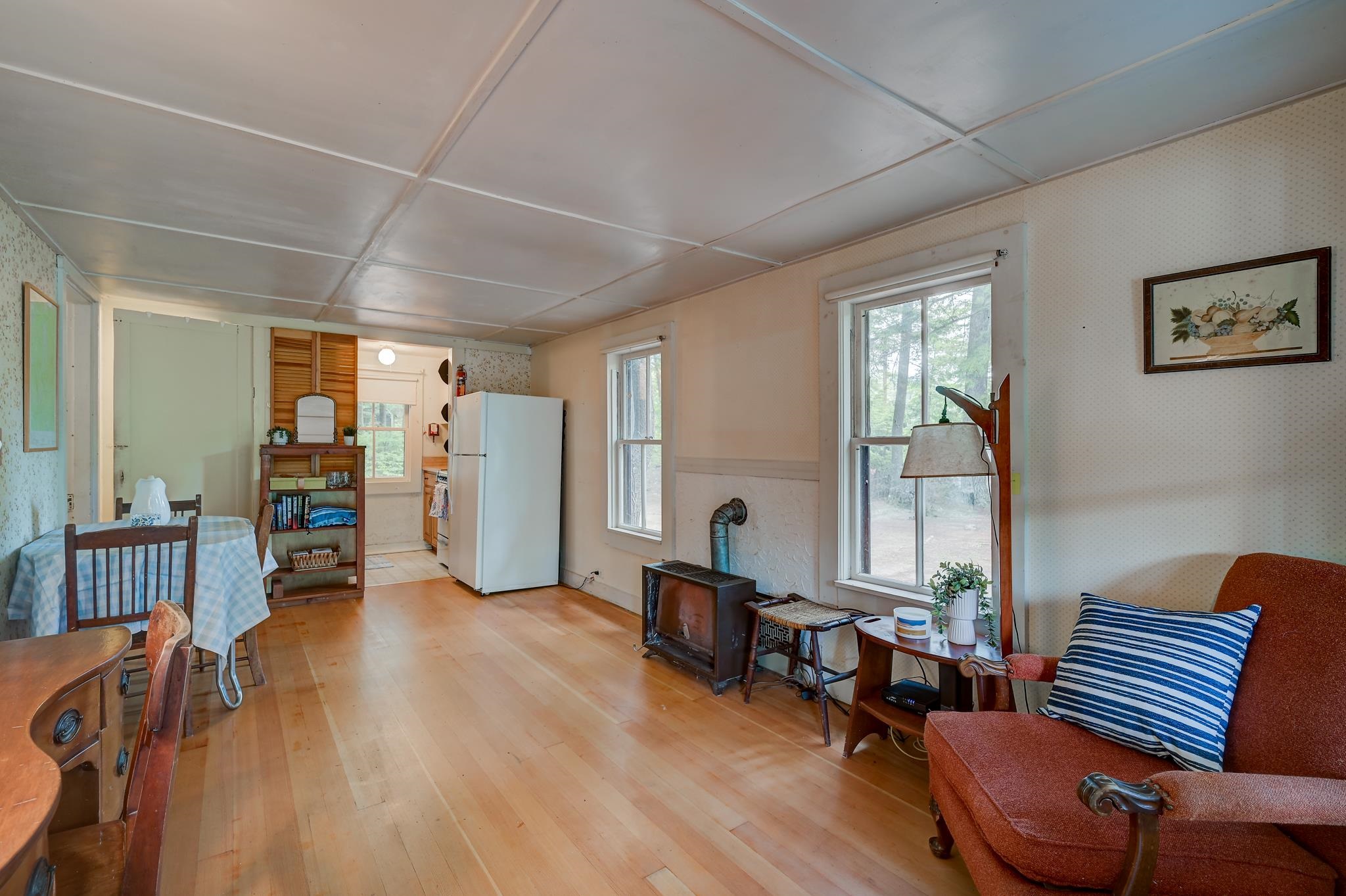 Living and dining area with a wood stove, light wood floors, and windows overlooking trees.