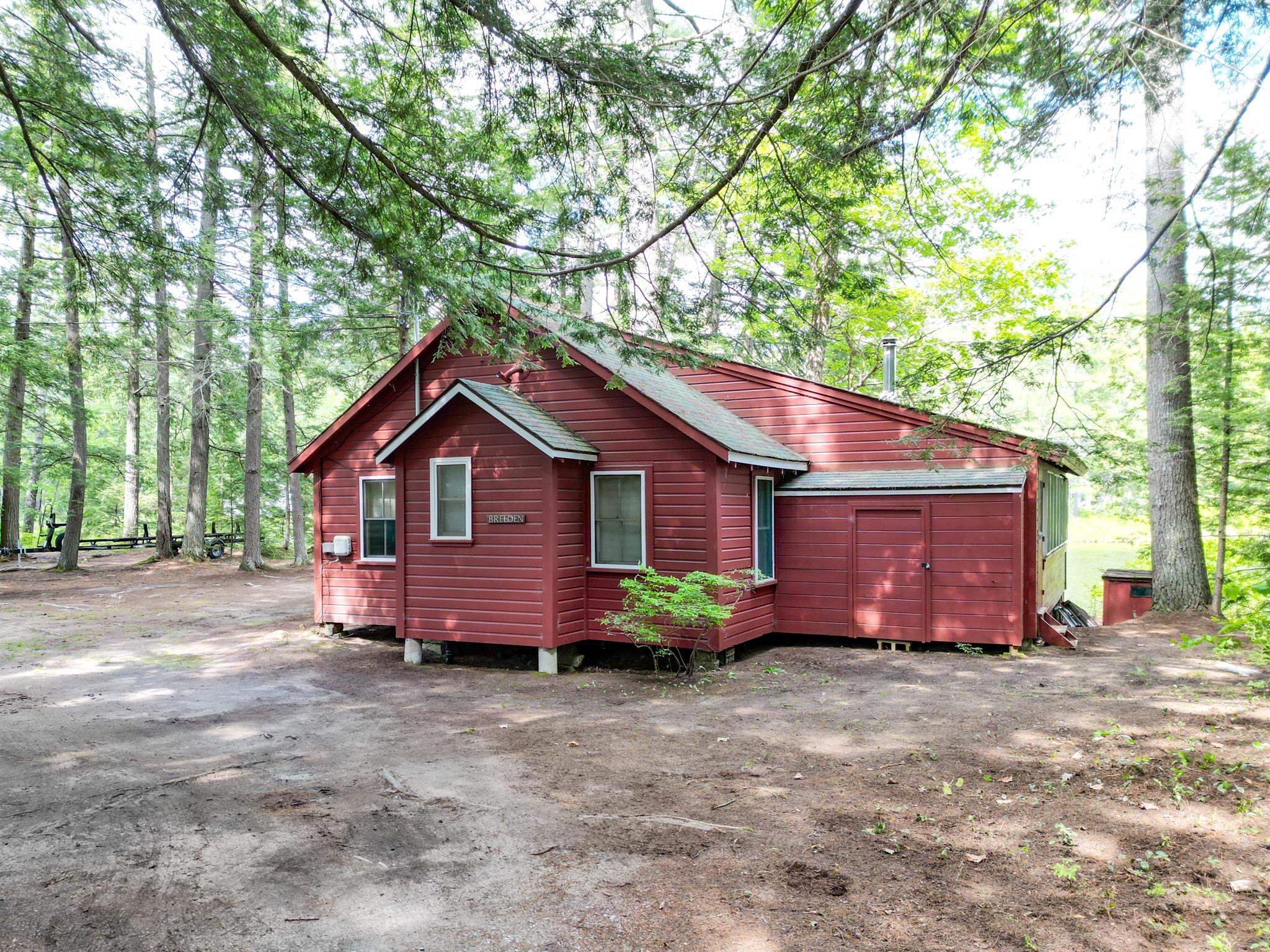 Red cottage nestled among mature trees, with a small attached garage and dirt driveway.