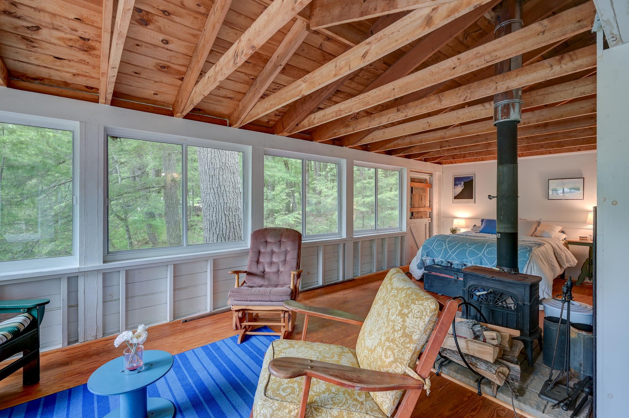 Bedroom and sitting area with exposed wood ceiling, forest-view windows, and a wood stove.