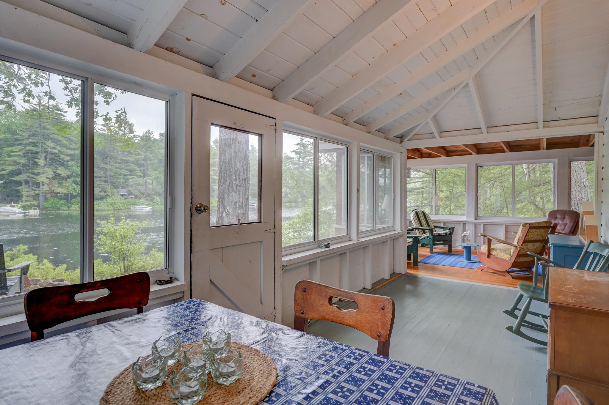 Enclosed porch with a dining table and sitting area overlooking a lake through wide windows.