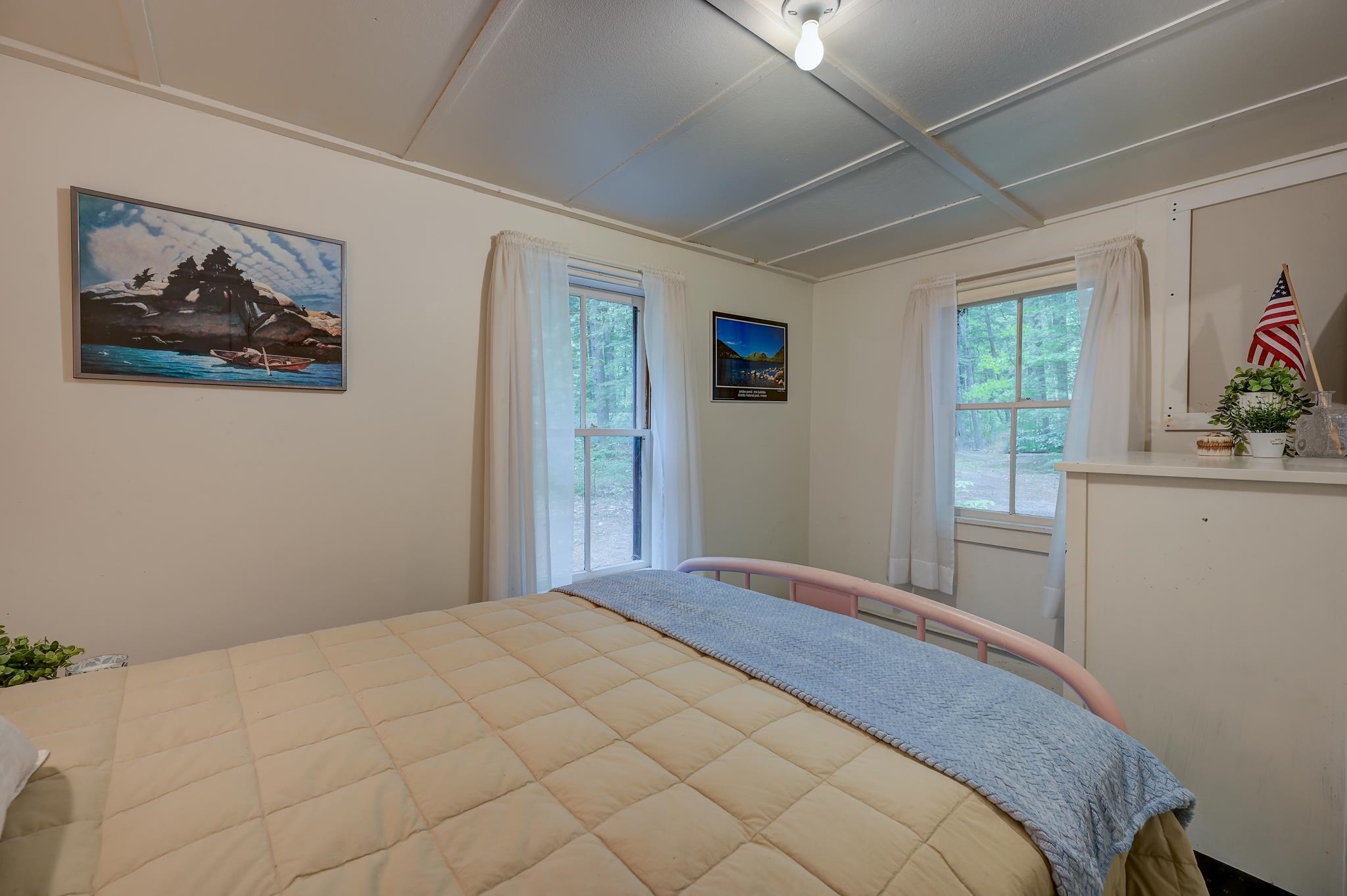 Bedroom with a large tan-quilted bed, blue throw, and two curtained windows overlooking trees.