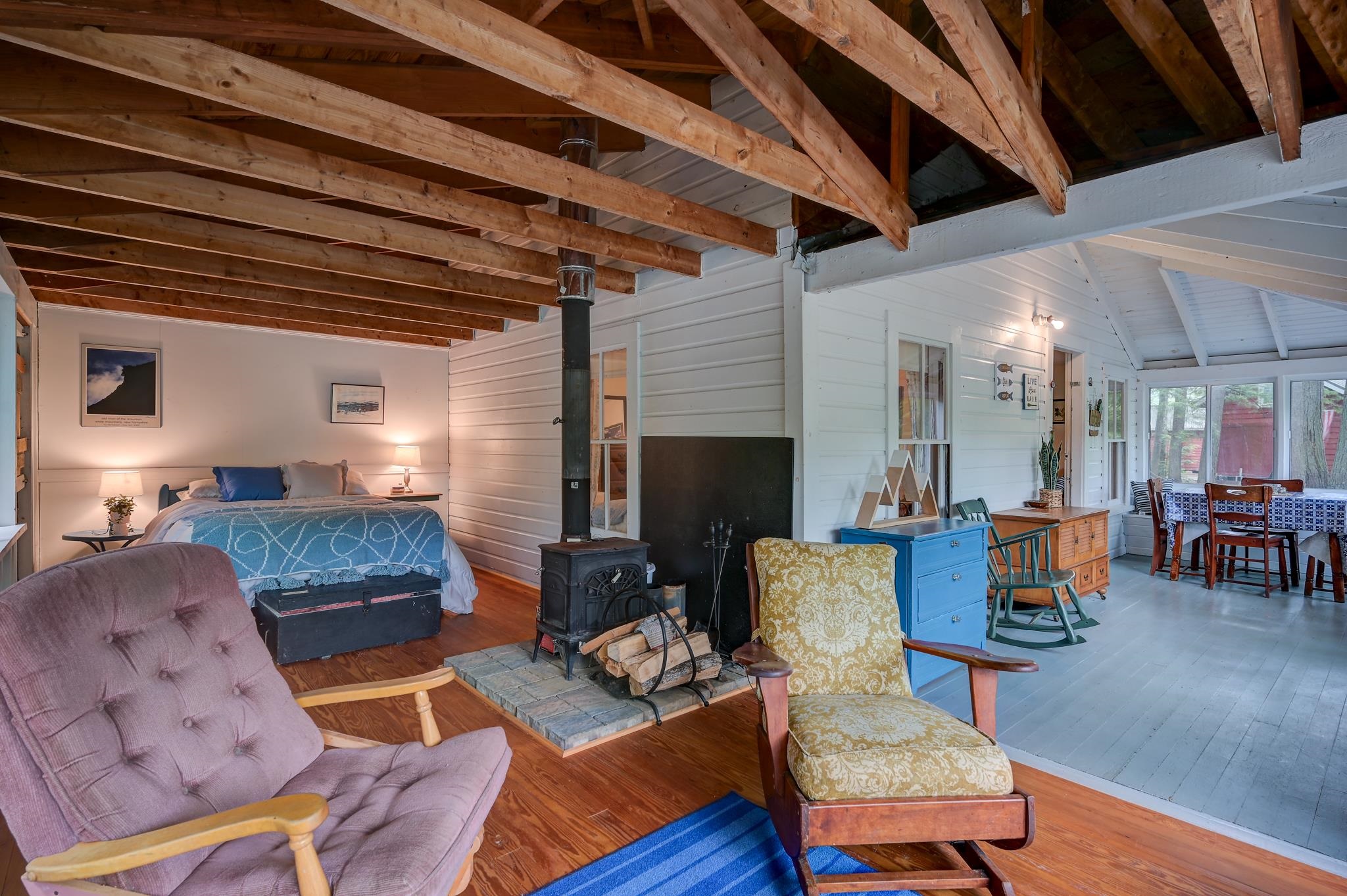 Open-plan bedroom and sitting area with exposed beams, a wood stove, and a dining nook by large windows.