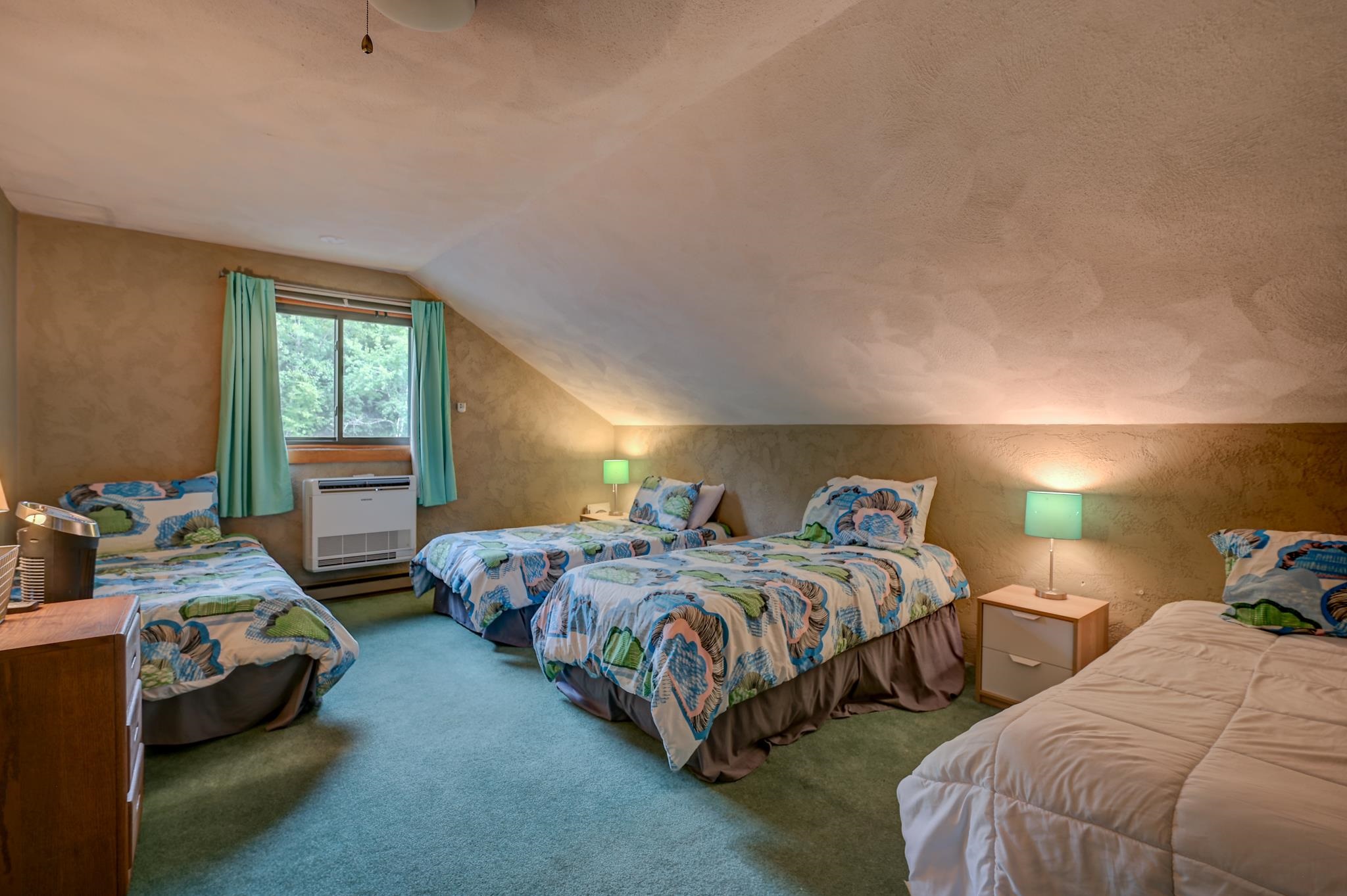 Attic bedroom with four beds, colorful patterned bedding, and a window framed by turquoise curtains.