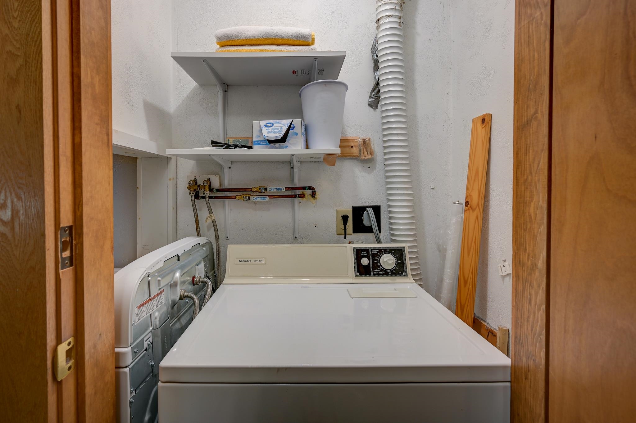 Laundry room with a top-loading washer, dryer, exposed utility hookups, and wall shelves.