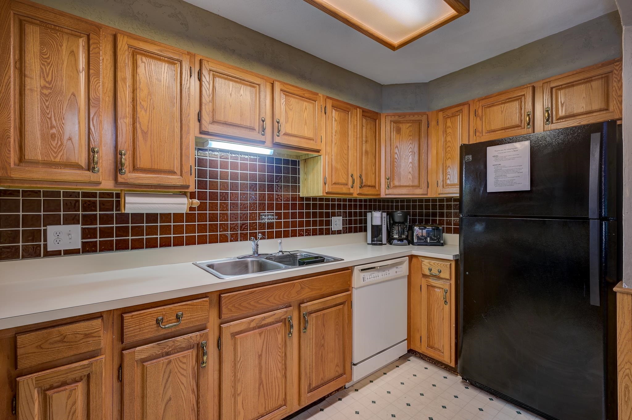 Kitchen with honey-colored cabinets, brown tile backsplash, double sink, and black refrigerator.