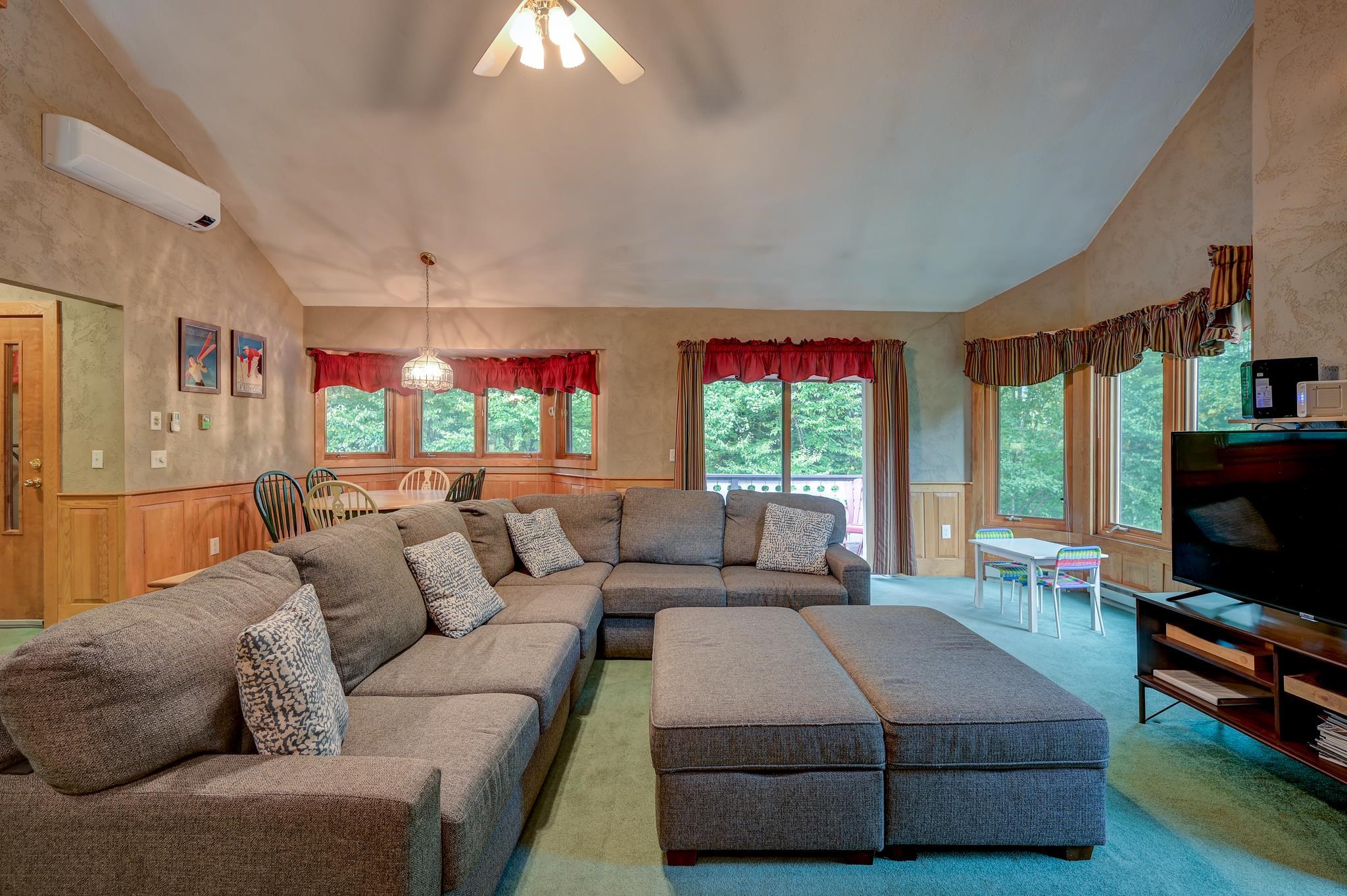Living room with a large gray sectional, ottoman, and wood-trimmed windows overlooking trees.