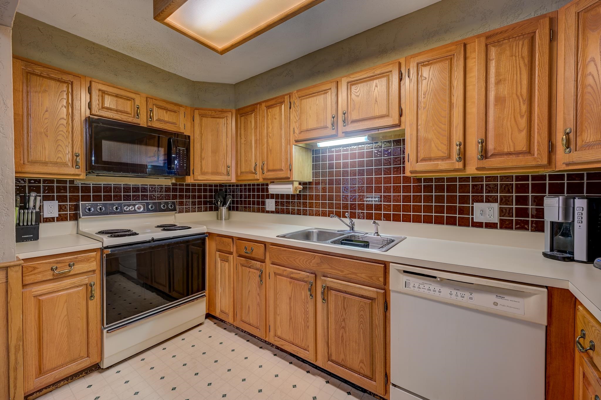 Kitchen with honey-toned wood cabinets, dark tile backsplash, white countertops, and electric range.