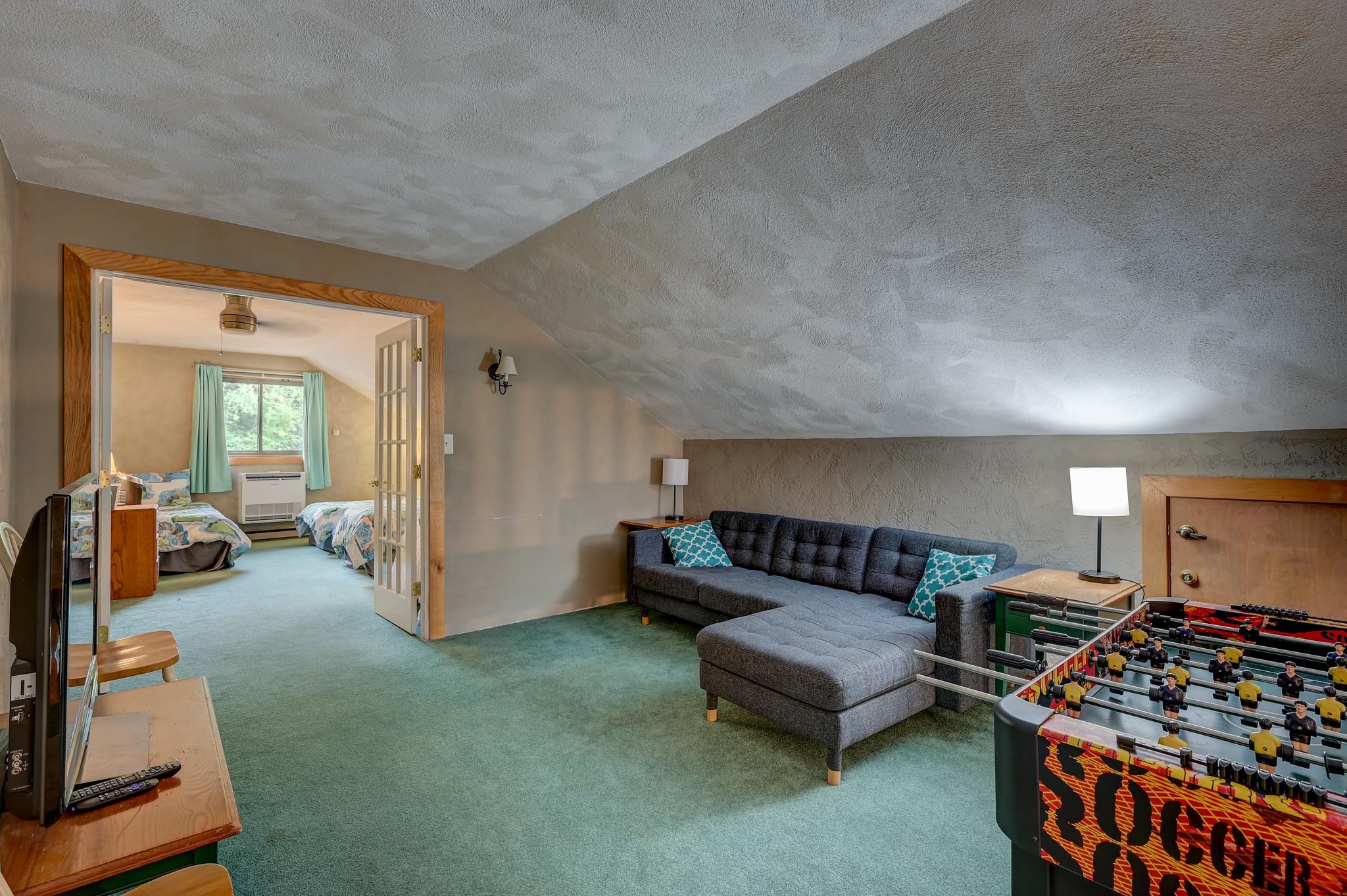 Attic loft with a gray sectional, foosball table, and doorway to a two-bed bedroom.