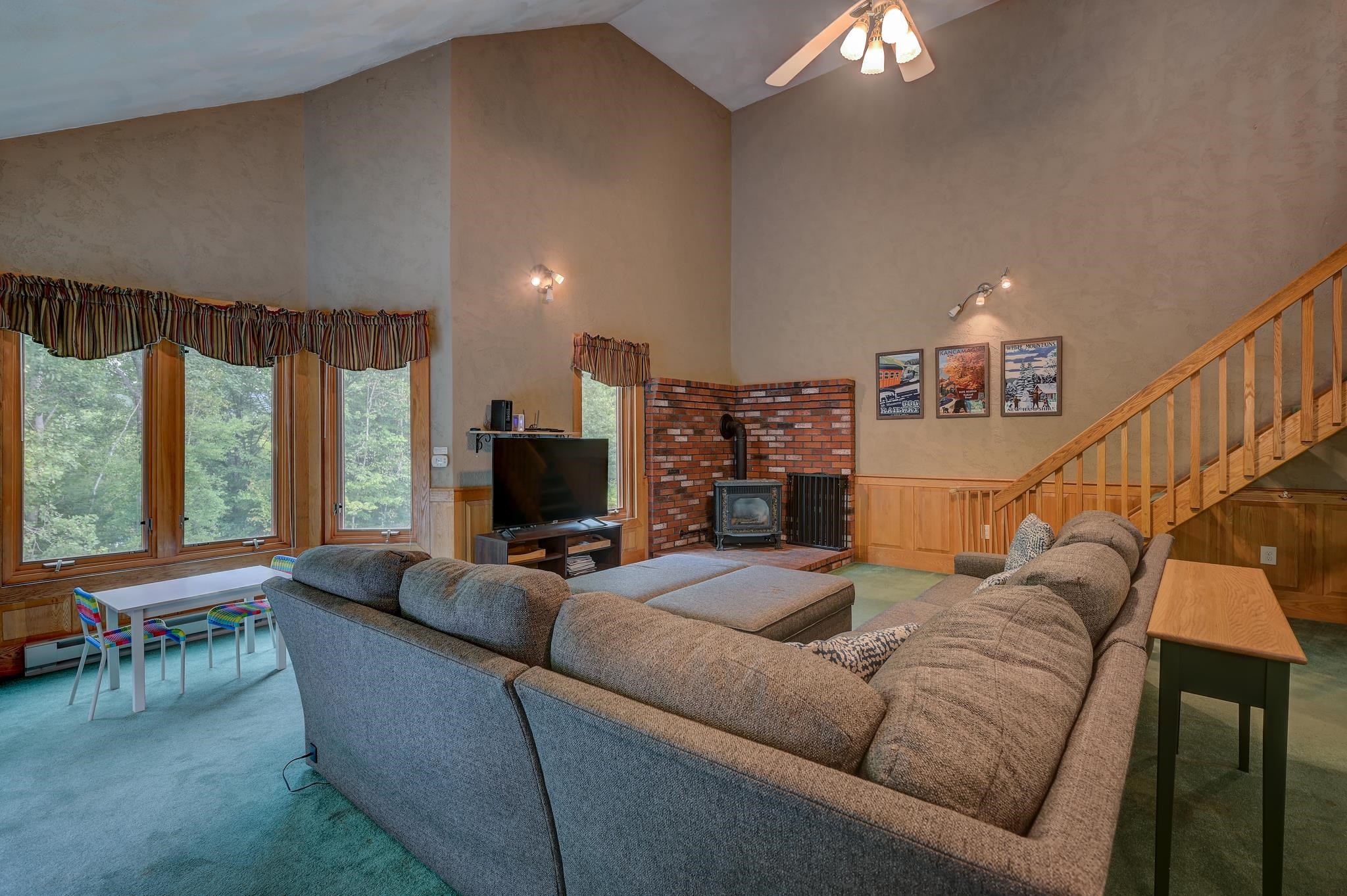 Living room with a vaulted ceiling, large windows, brick fireplace and wood stove, sectional sofa, and staircase.