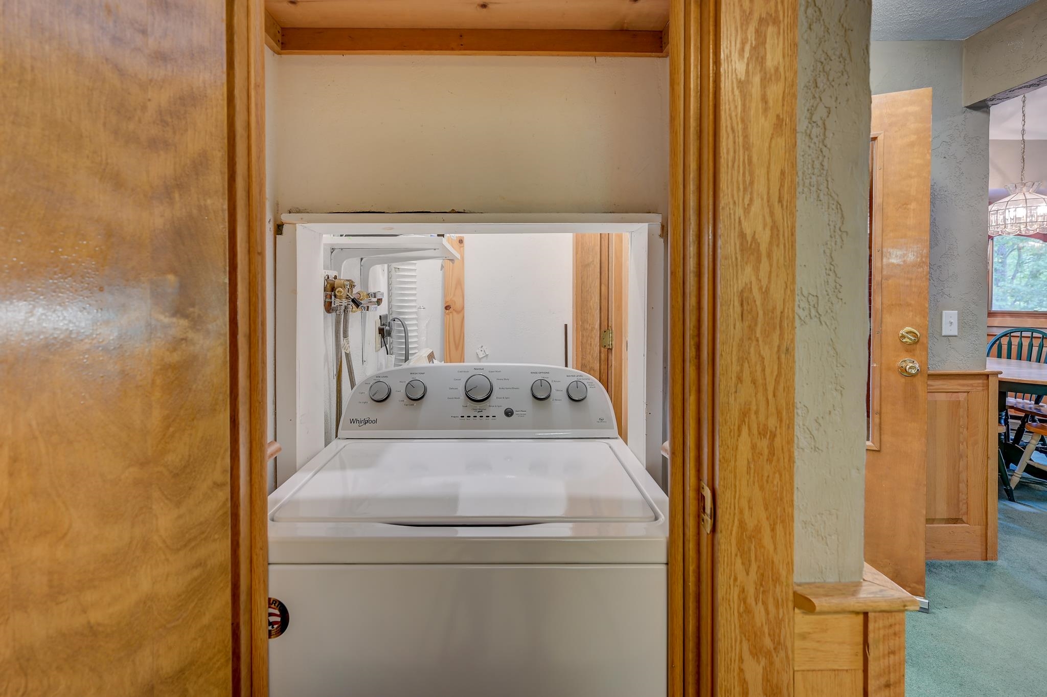 Top-loading washer in a wooden laundry closet, with a dining area visible through the doorway.