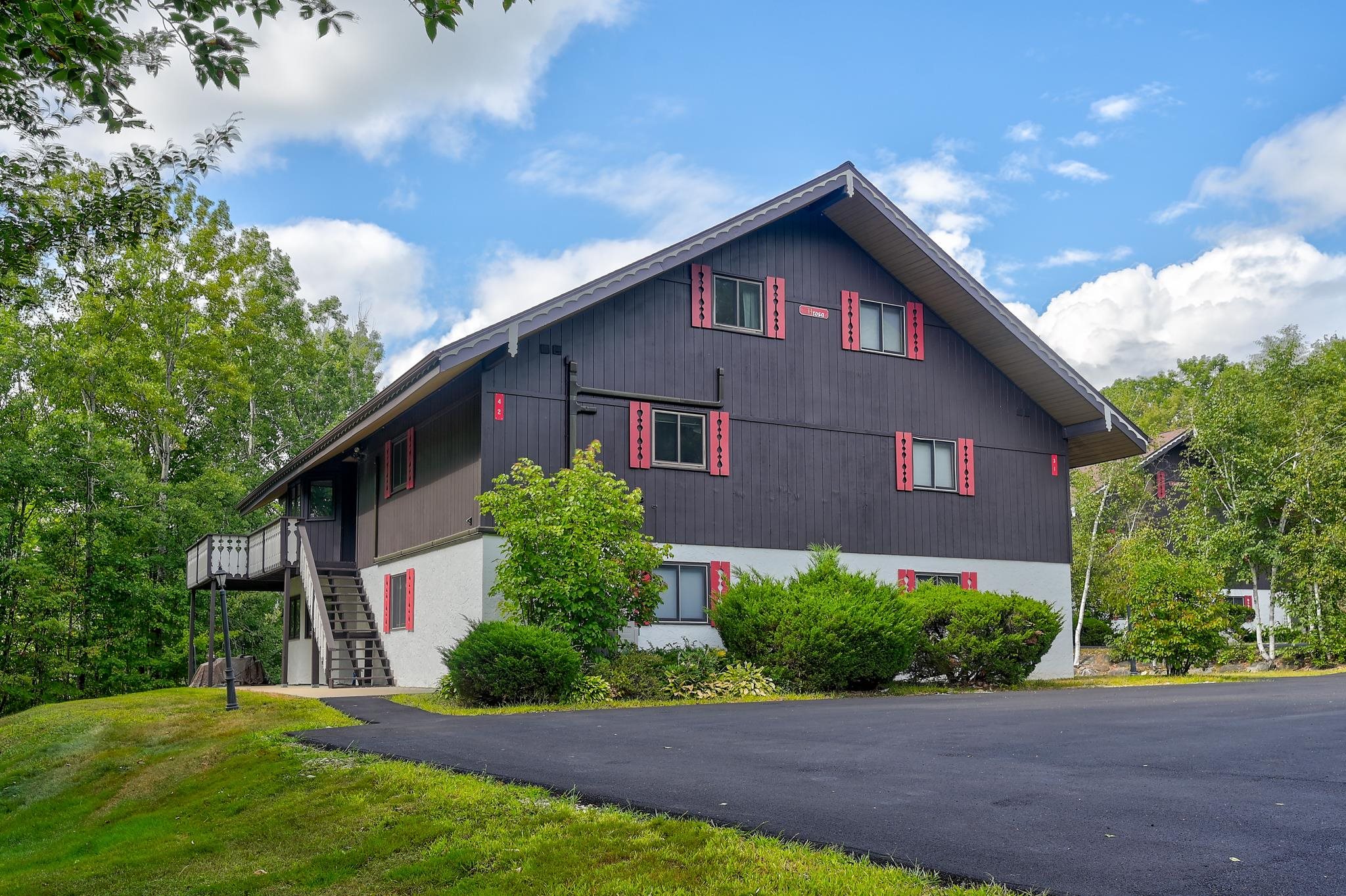 Multi-unit building with dark siding, red shutters, and an elevated deck beside a paved parking area.