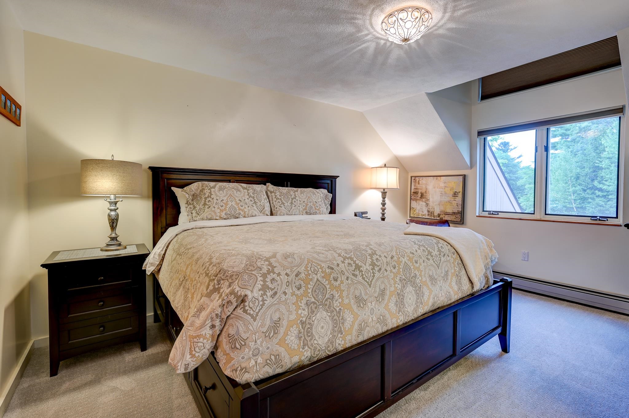 Furnished bedroom with a dark wood bed, patterned cream bedding, and a wide window overlooking trees.