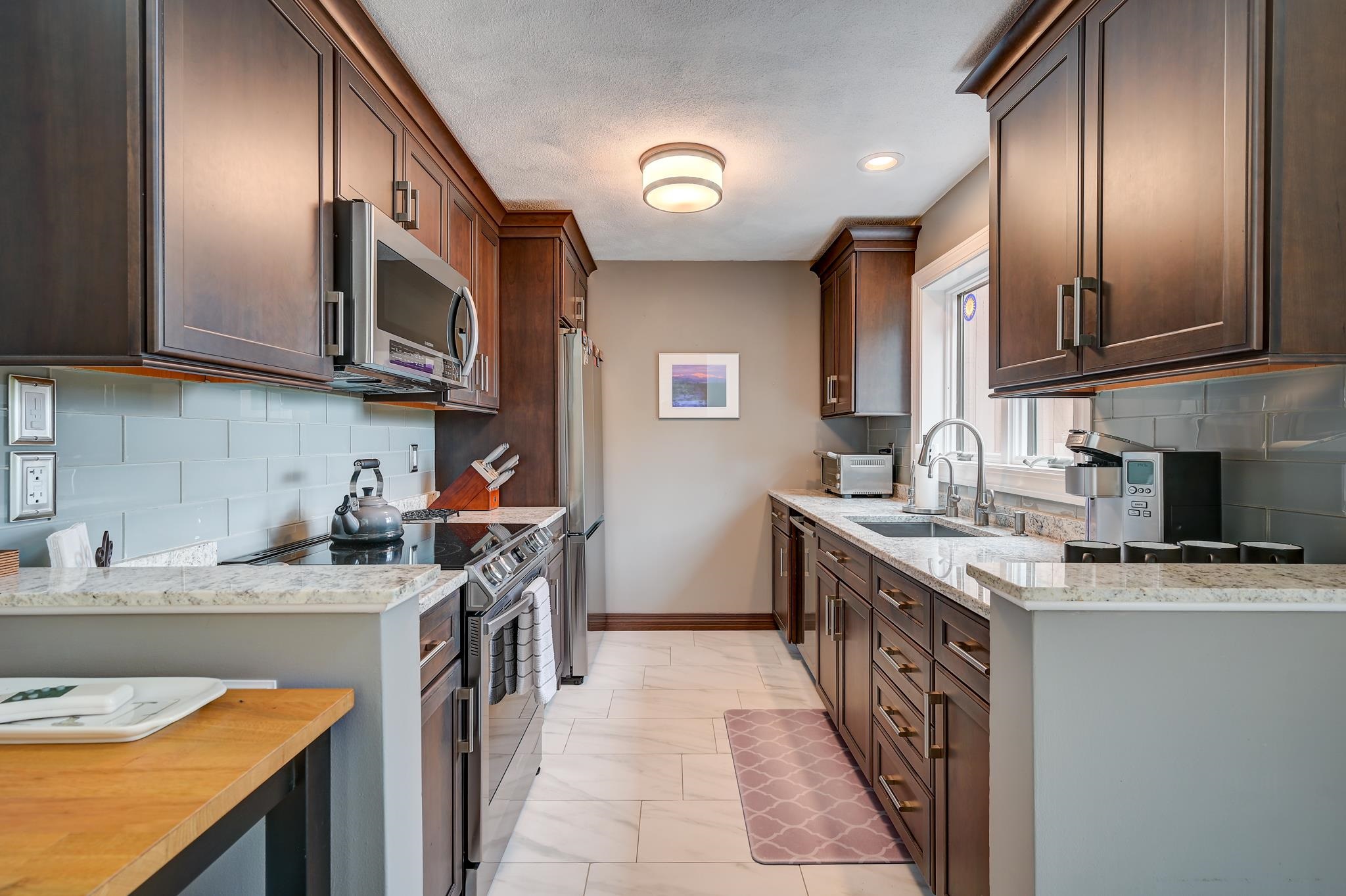 Galley kitchen with dark wood cabinetry, pale stone counters, and a double sink beneath the window.