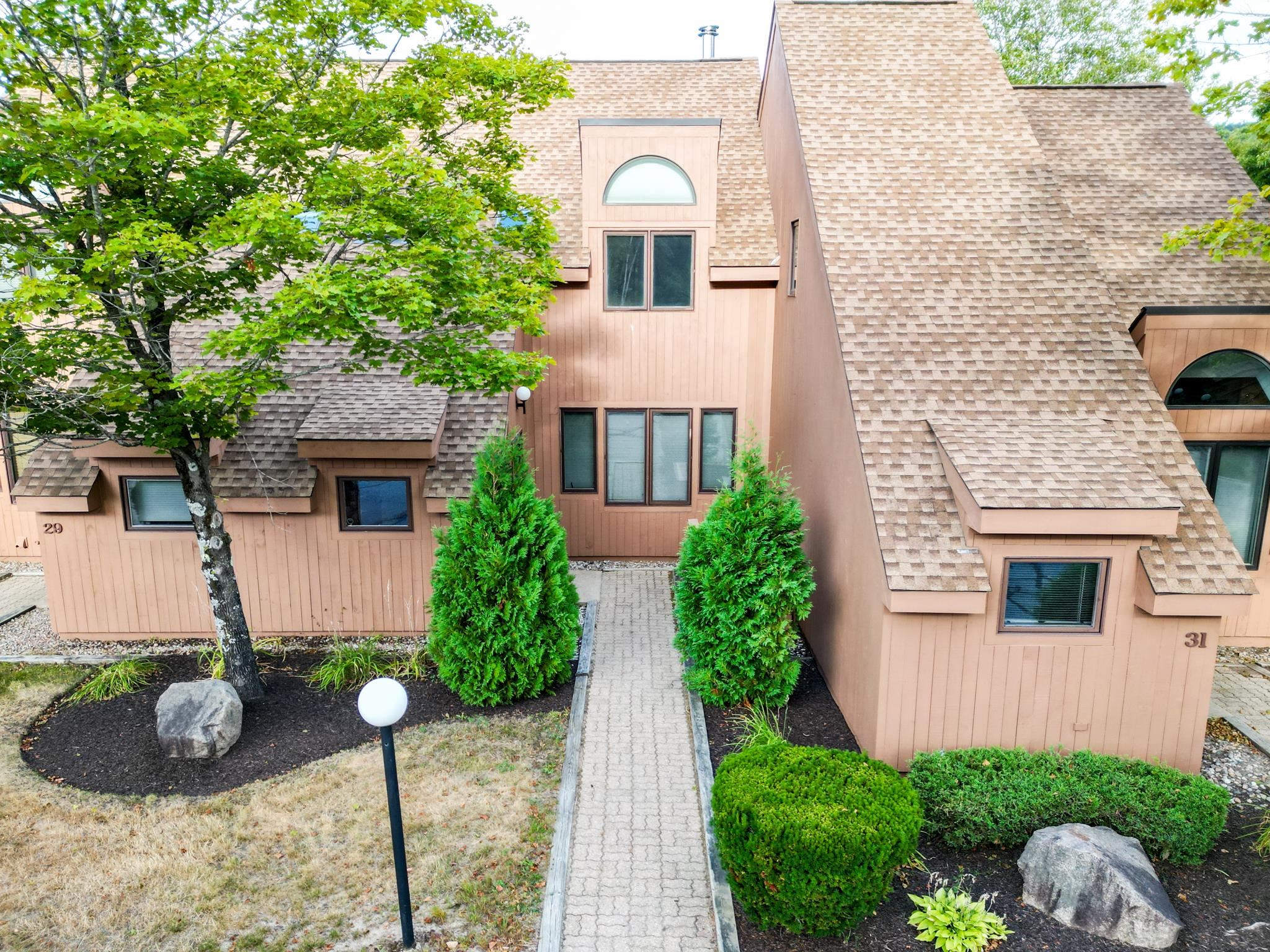 Attached brown townhomes with steep roofs flank a paved walkway bordered by trees and shrubs.