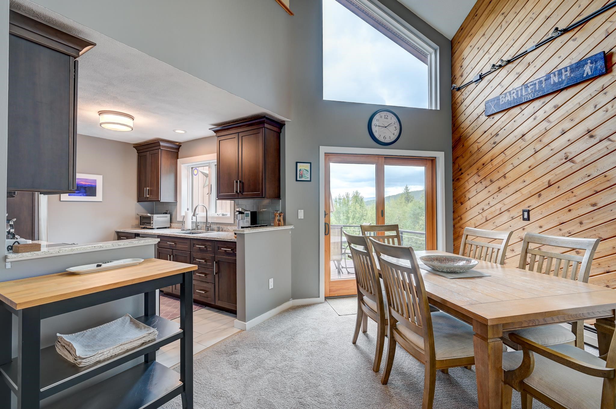 Open kitchen and dining area with vaulted ceiling, wooden table, and balcony doors overlooking trees and hills.