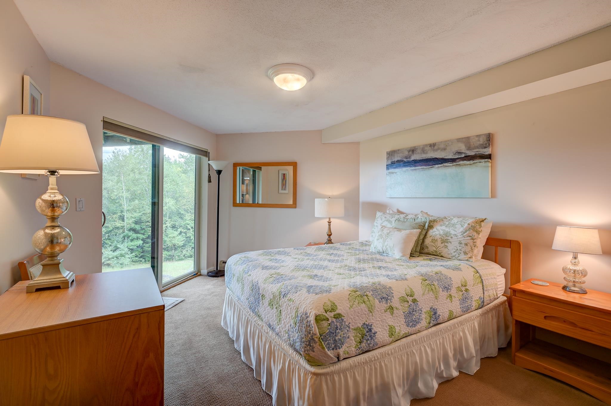 Furnished bedroom with a floral quilted bed beside a sliding glass door overlooking greenery.
