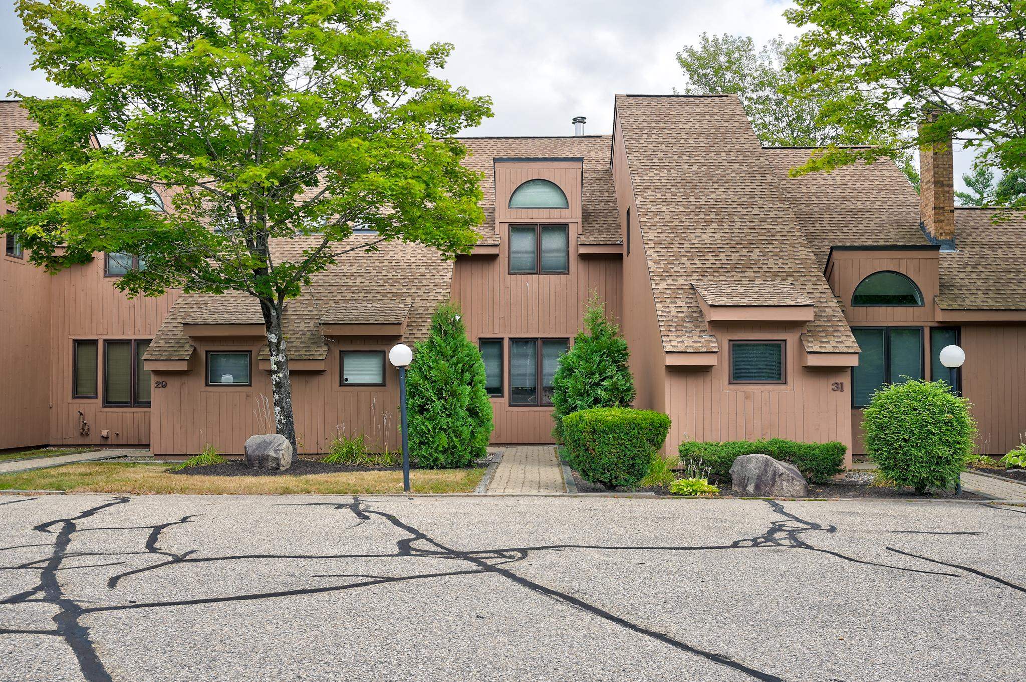 Brown condominium building with steep shingle roofs, arched windows, landscaped shrubs, and a paved parking area.