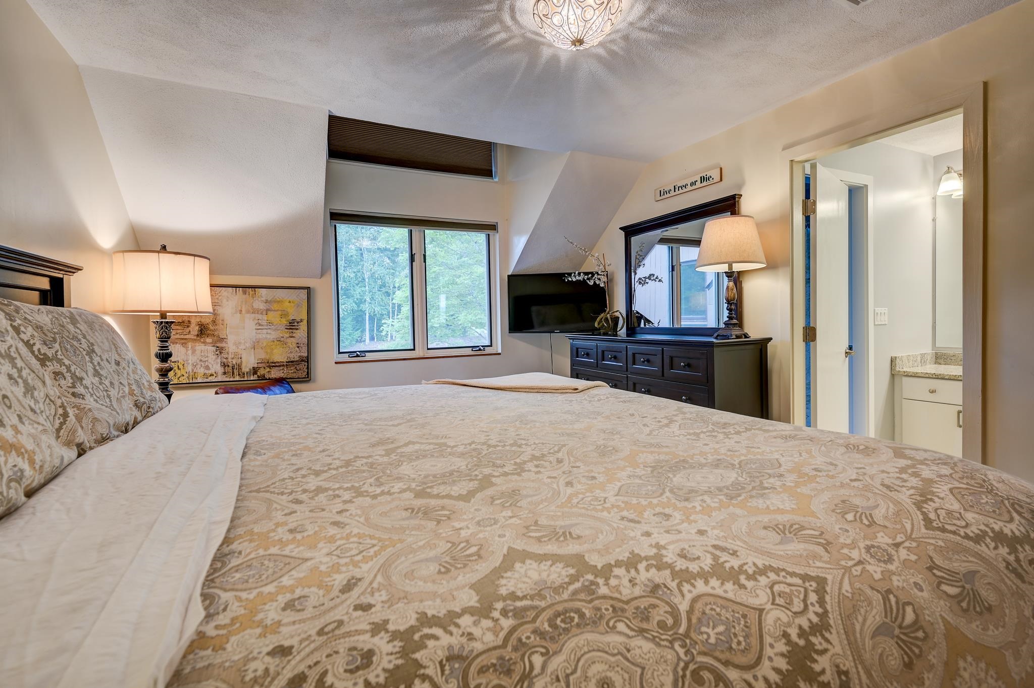 Furnished bedroom with a patterned bed, dark dresser, and windows overlooking trees.