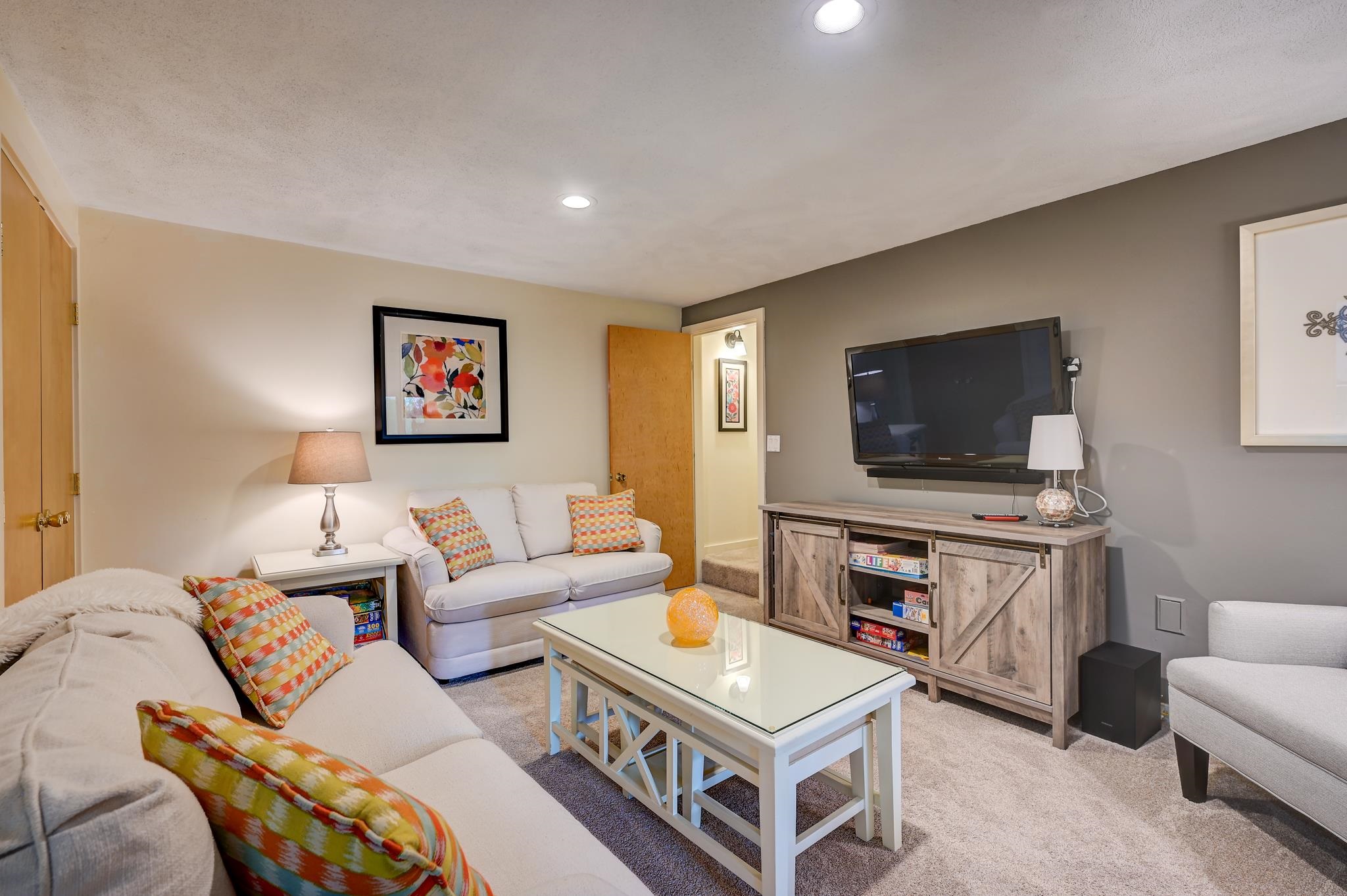 Living room with light sofas, colorful patterned cushions, and a wall-mounted television.
