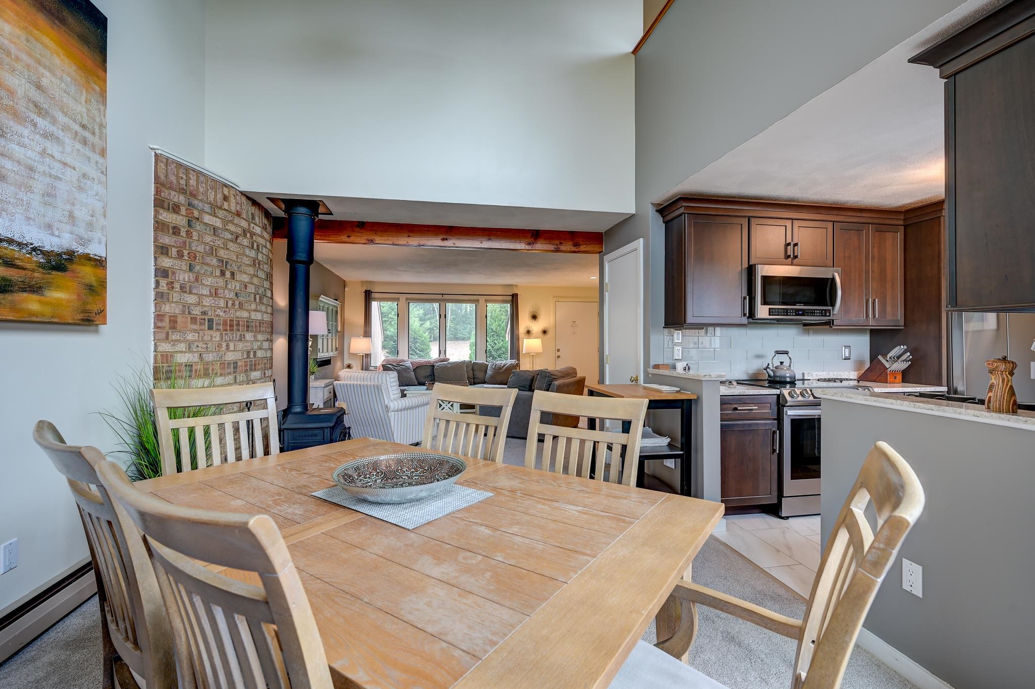Open dining and living area with a wood table, dark kitchen cabinets, brick wall, and wood stove.