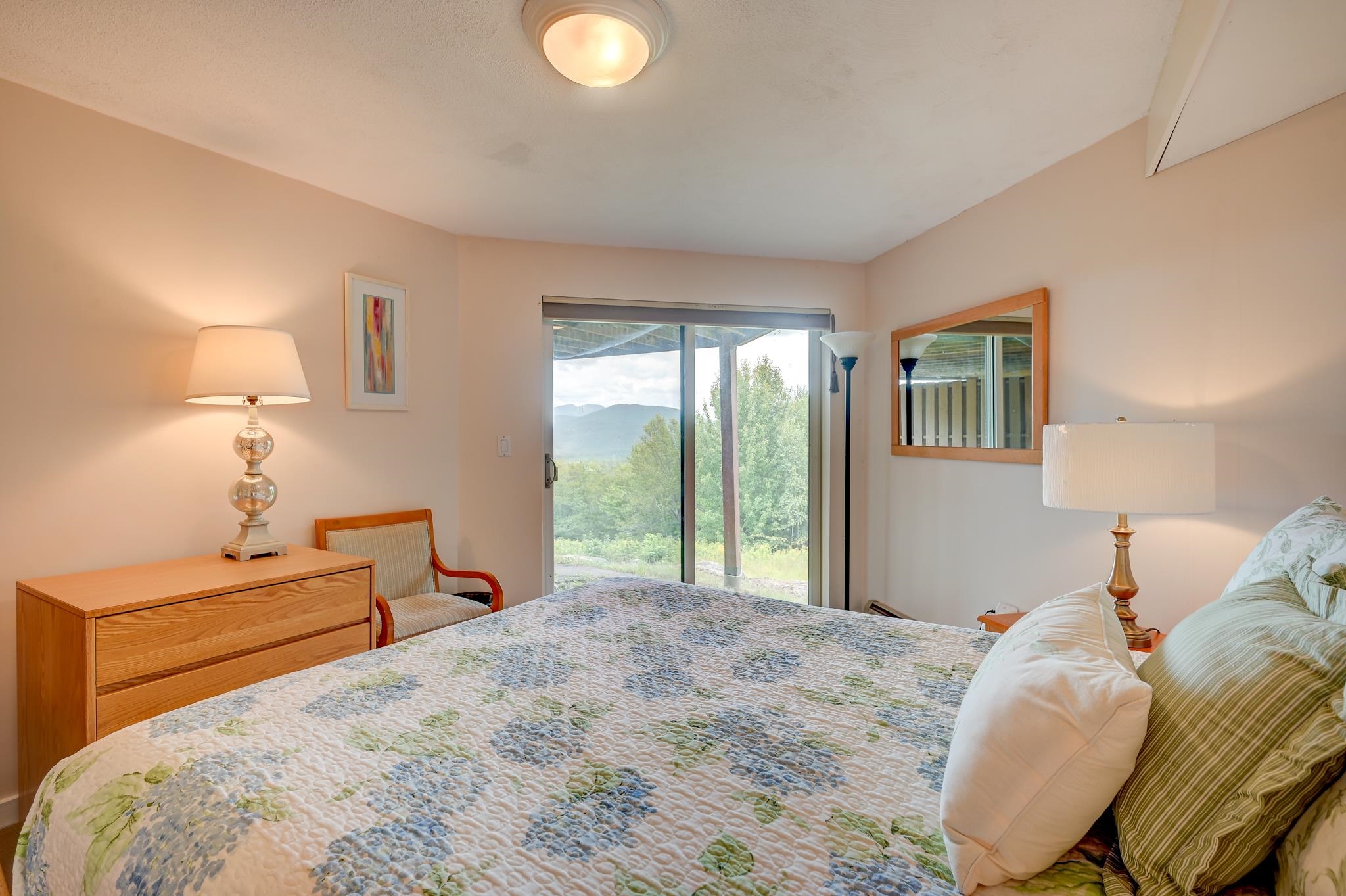 Bedroom with a floral quilt and sliding glass door overlooking trees and distant mountains.