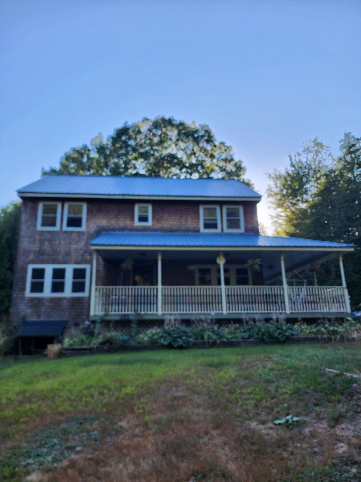 Two-story shingle house with a blue metal roof, long covered porch, and lawn.