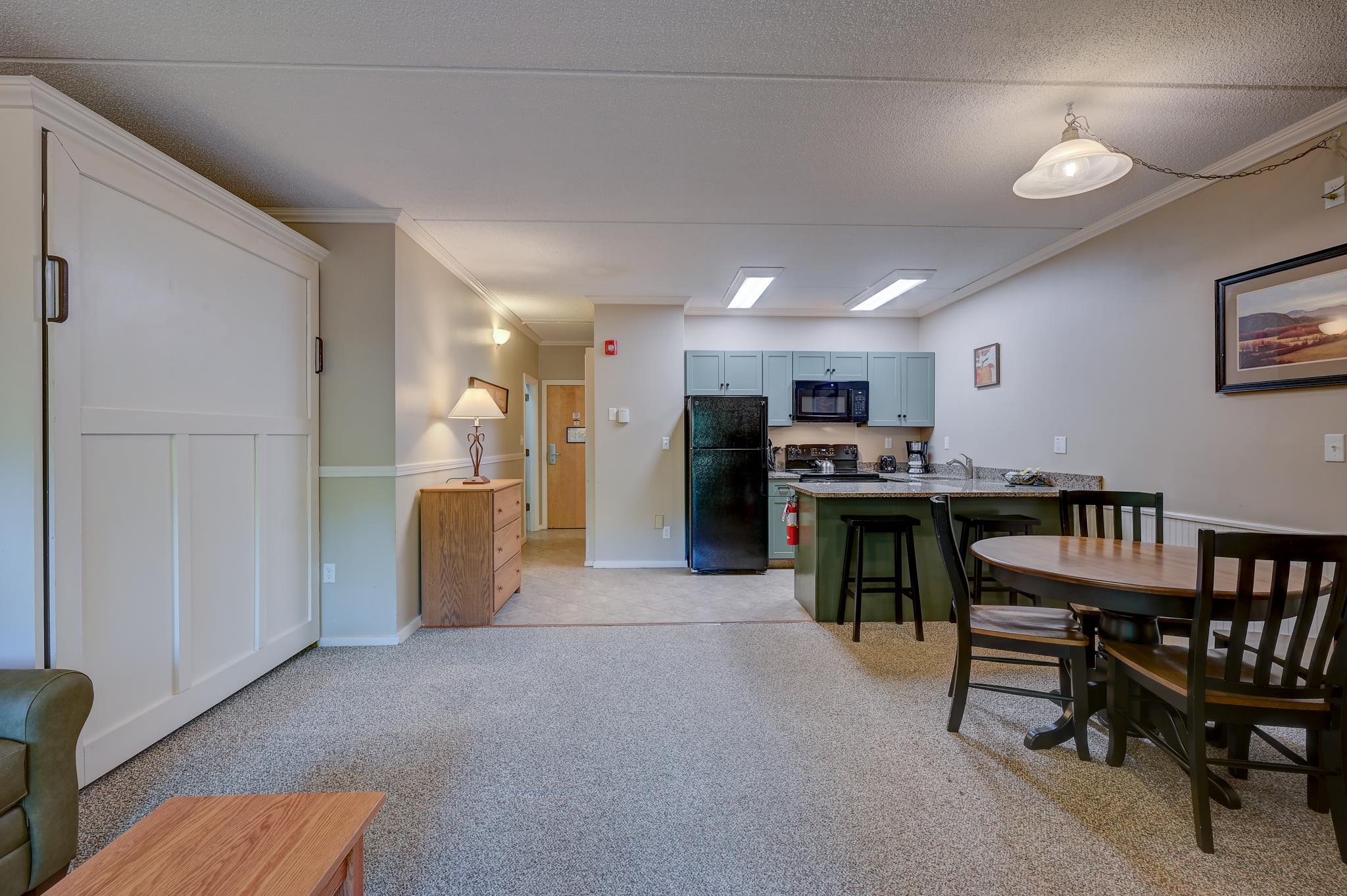 Open-plan condo living area with a cabinet bed, dining table, and compact kitchen with blue cabinets.