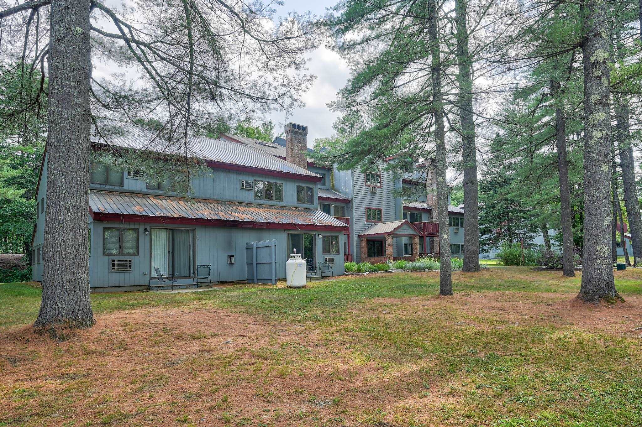 Blue-gray condominium building among tall pine trees, with a small patio and metal roofs.