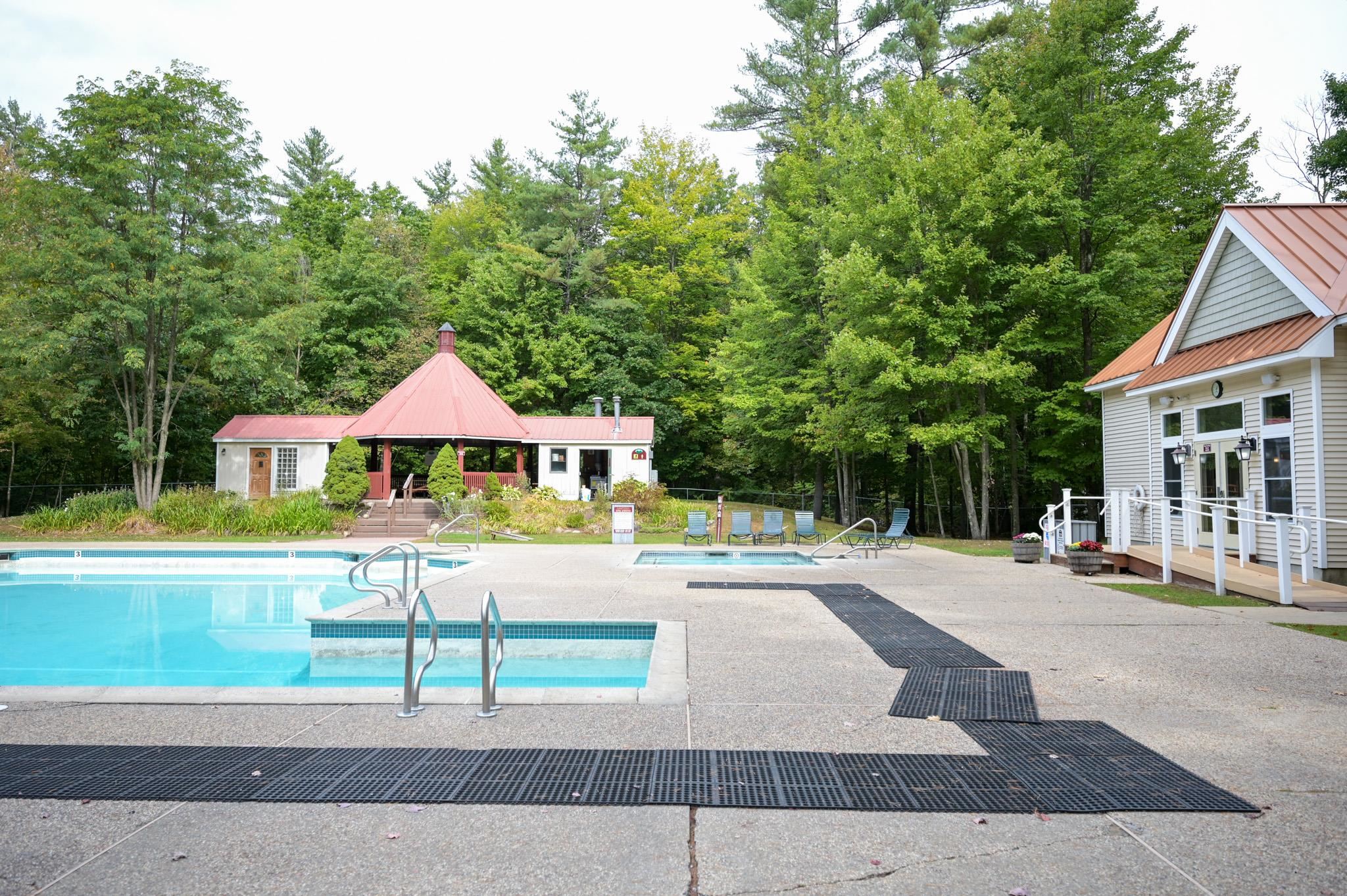 Community pool with a shallow section, red-roofed pavilion, lounge chairs, and wooded surroundings.