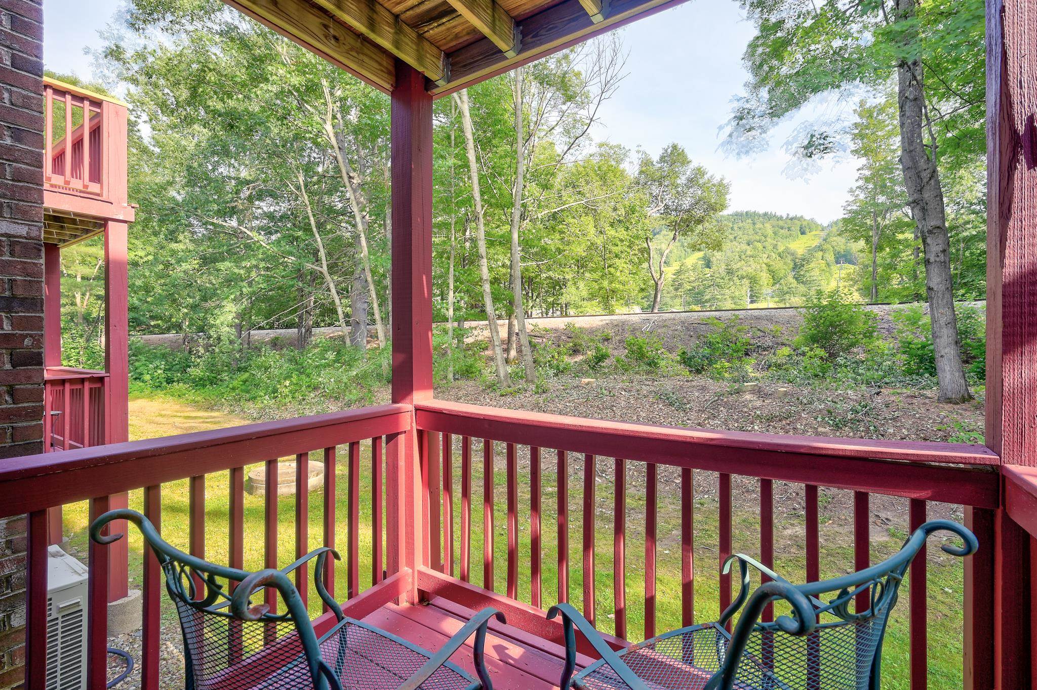 Covered balcony with red railings and two metal chairs overlooking a wooded hillside.