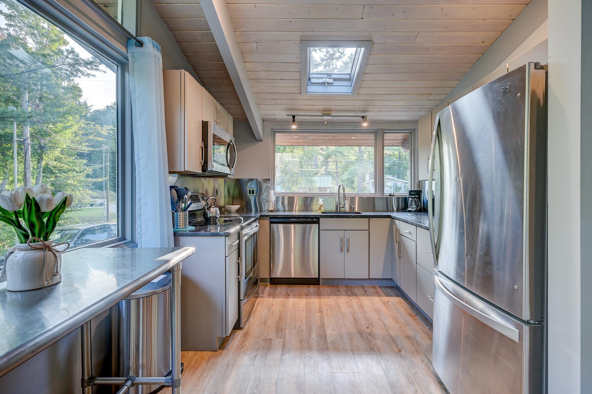 Modern kitchen with stainless steel appliances, large windows, and a wood-plank ceiling.